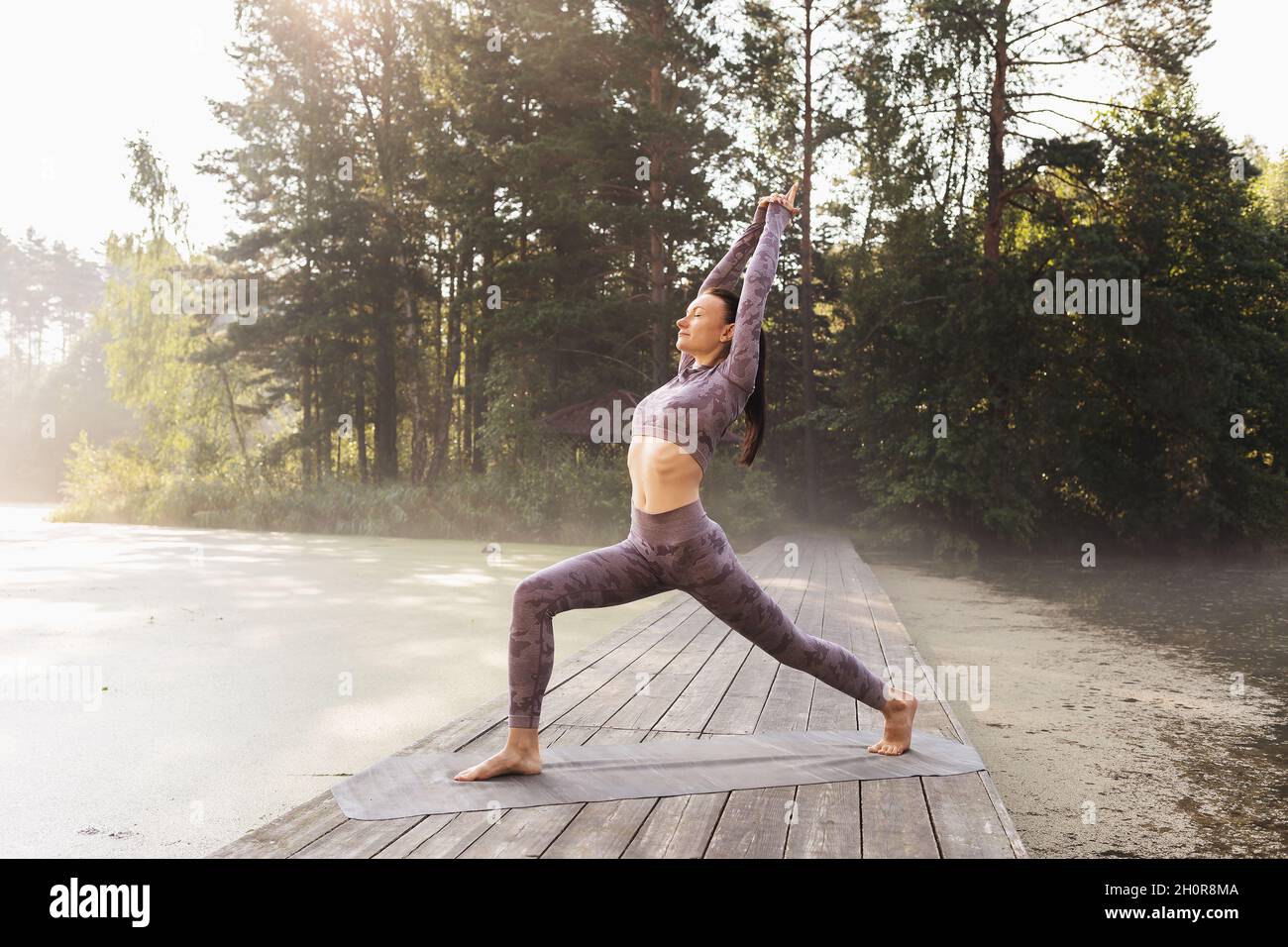 Une femme exécute l'exercice de virabhadrasana, la première posture de guerrier, pratique le yoga, se tient le matin sur un pont en bois dans le parc Banque D'Images