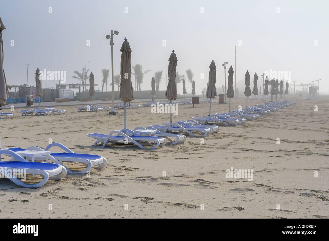 Plage brumeuse avec des rangées floues de parasols fermés, lit de soleil en dehors de la saison le jour brumeux.brouillard épais sur la plage, chaises longues vides, parasols fermés. Banque D'Images