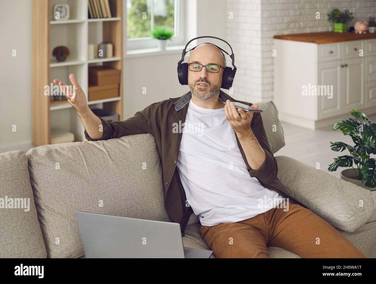 Homme enregistrant un message vocal ou parlant sur un haut-parleur lorsqu'il est assis sur un canapé à la maison Banque D'Images