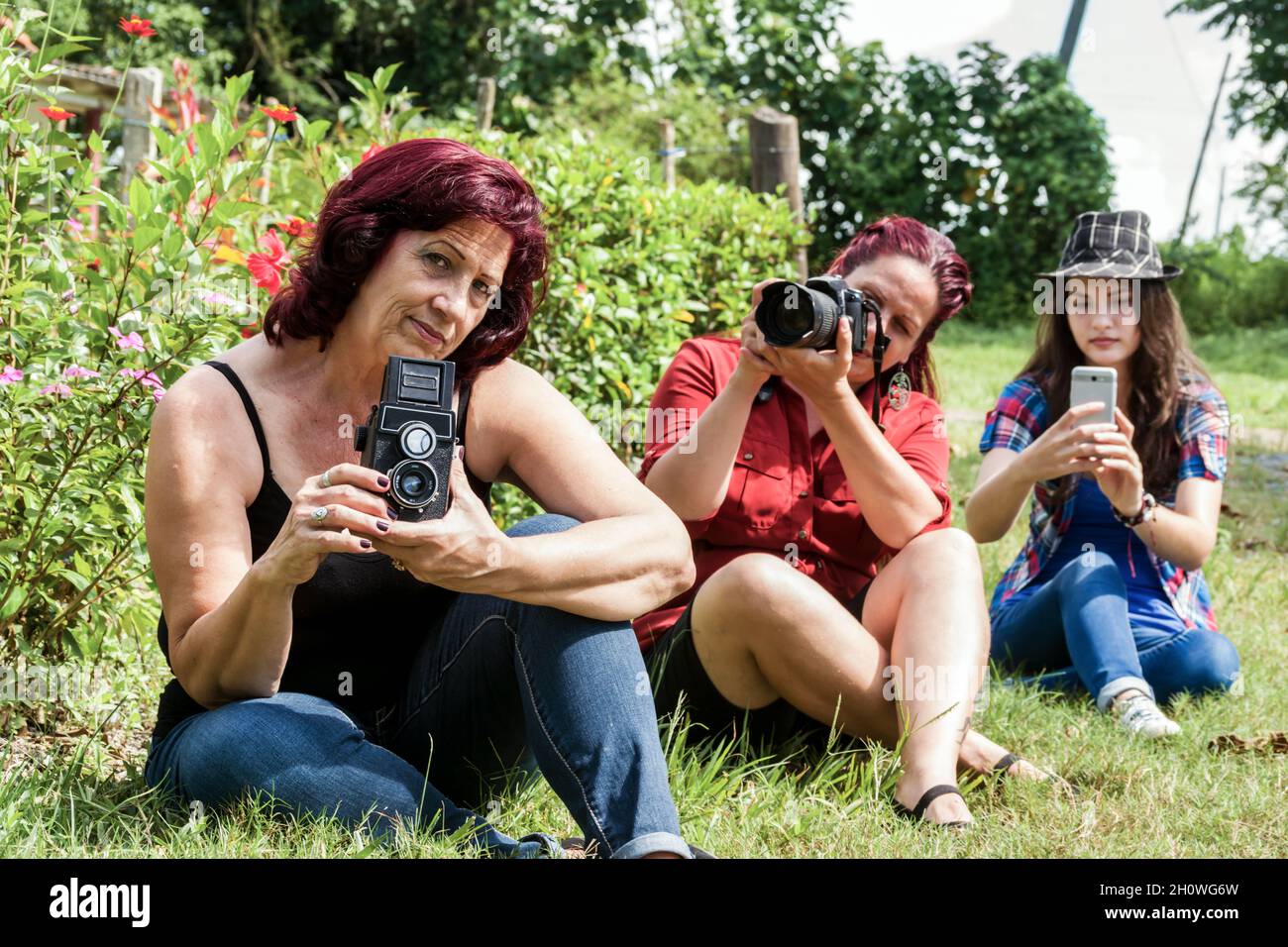 Différentes générations de femmes latines à l'extérieur, assis sur l'herbe, chacune d'entre elles tient une caméra qui correspond à sa génération Banque D'Images