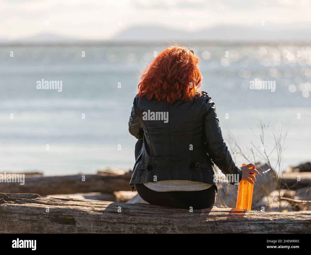 Vue arrière d'une jeune femme assise sur la plage et regardant la mer avec une bouteille d'eau à la main.Méditation dans la nature. vue sur la rue, photo de voyage, Banque D'Images