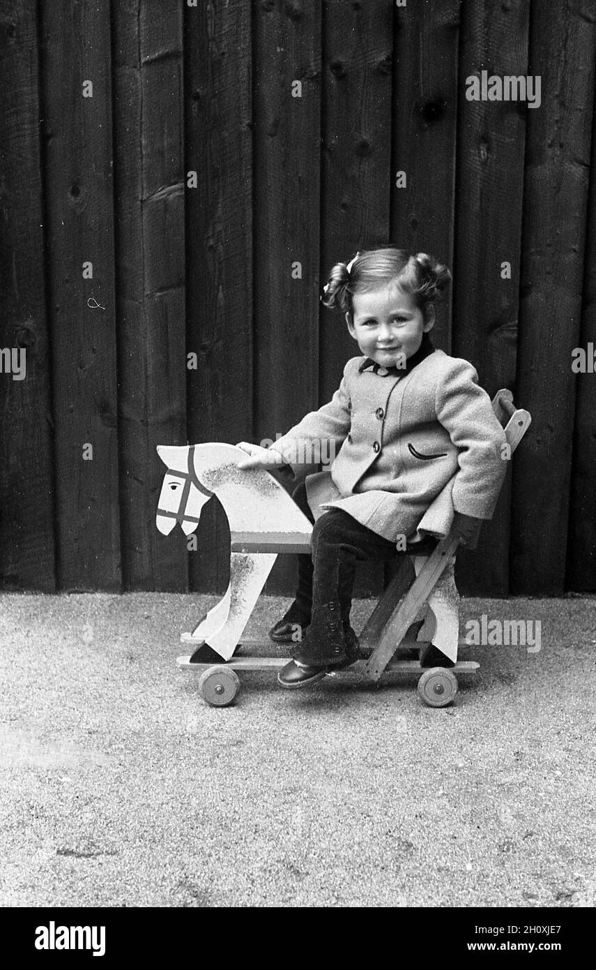 Années 1960, historique, à l'extérieur sur une zone de gravier plat, une petite fille dans un manteau assis sur son jouet porté, une poussée de bois le long de cheval sur roues, Angleterre, Royaume-Uni. Banque D'Images