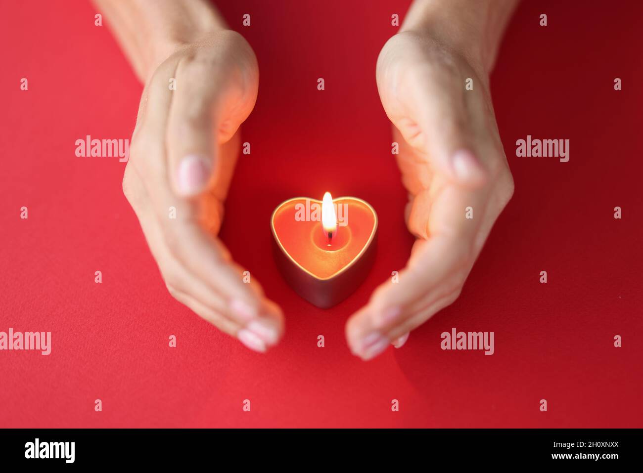 La main femelle protège soigneusement la bougie brûlante en forme de coeur sur fond rouge Banque D'Images