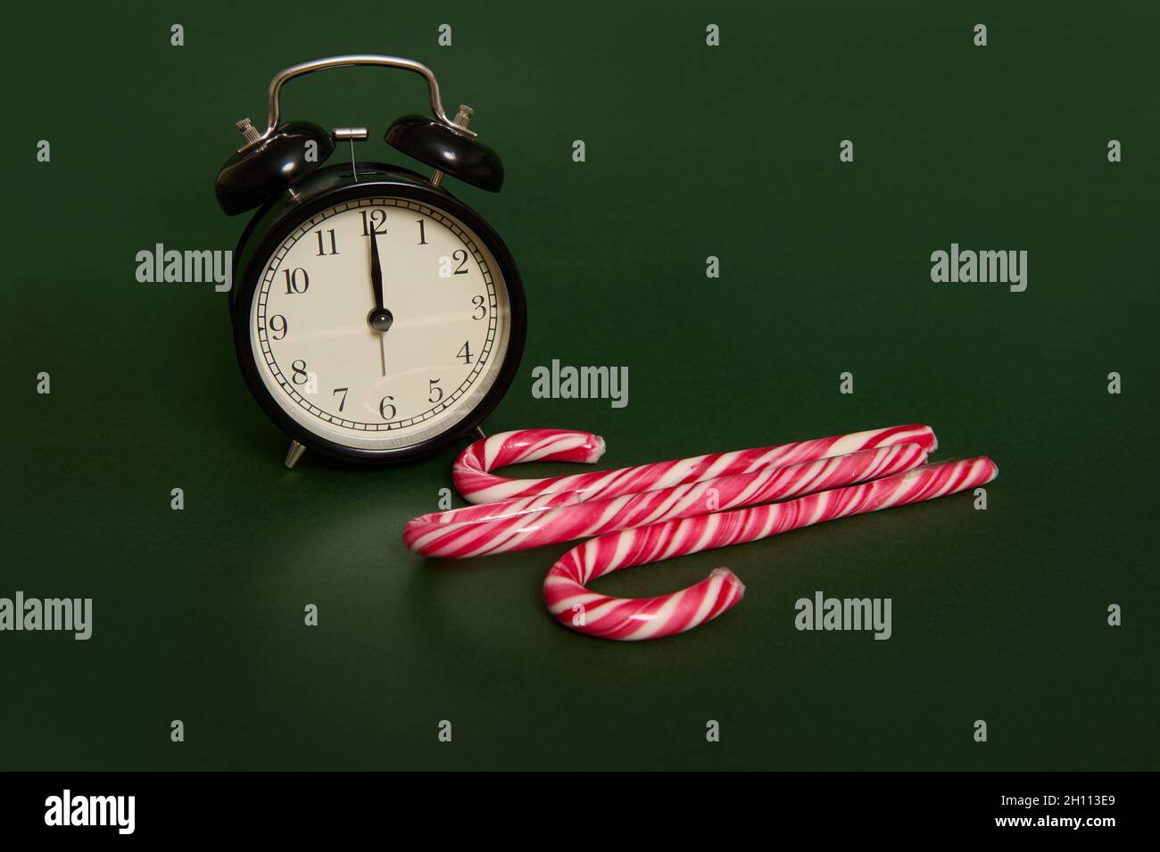 Studio photographié avec des ombres d'un réveil noir avec minuit sur le cadran de l'horloge et des sucettes de Noël à rayures sucrées, des cannes à sucre isolées sur le noir Banque D'Images