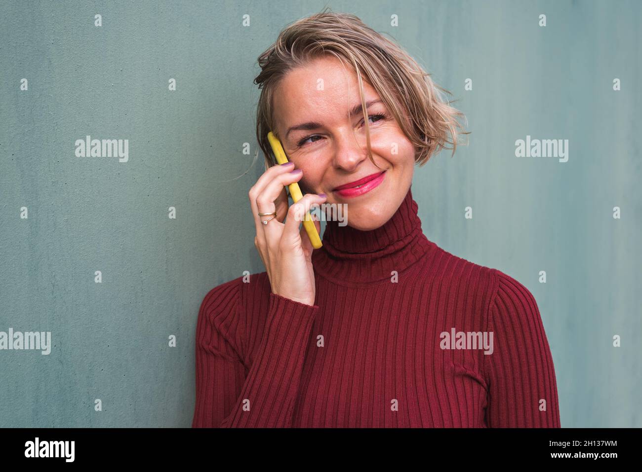 portrait horizontal d'une femme blonde sur un chandail en grenat parlant au téléphone tout en s'appuyant sur un mur vert Banque D'Images