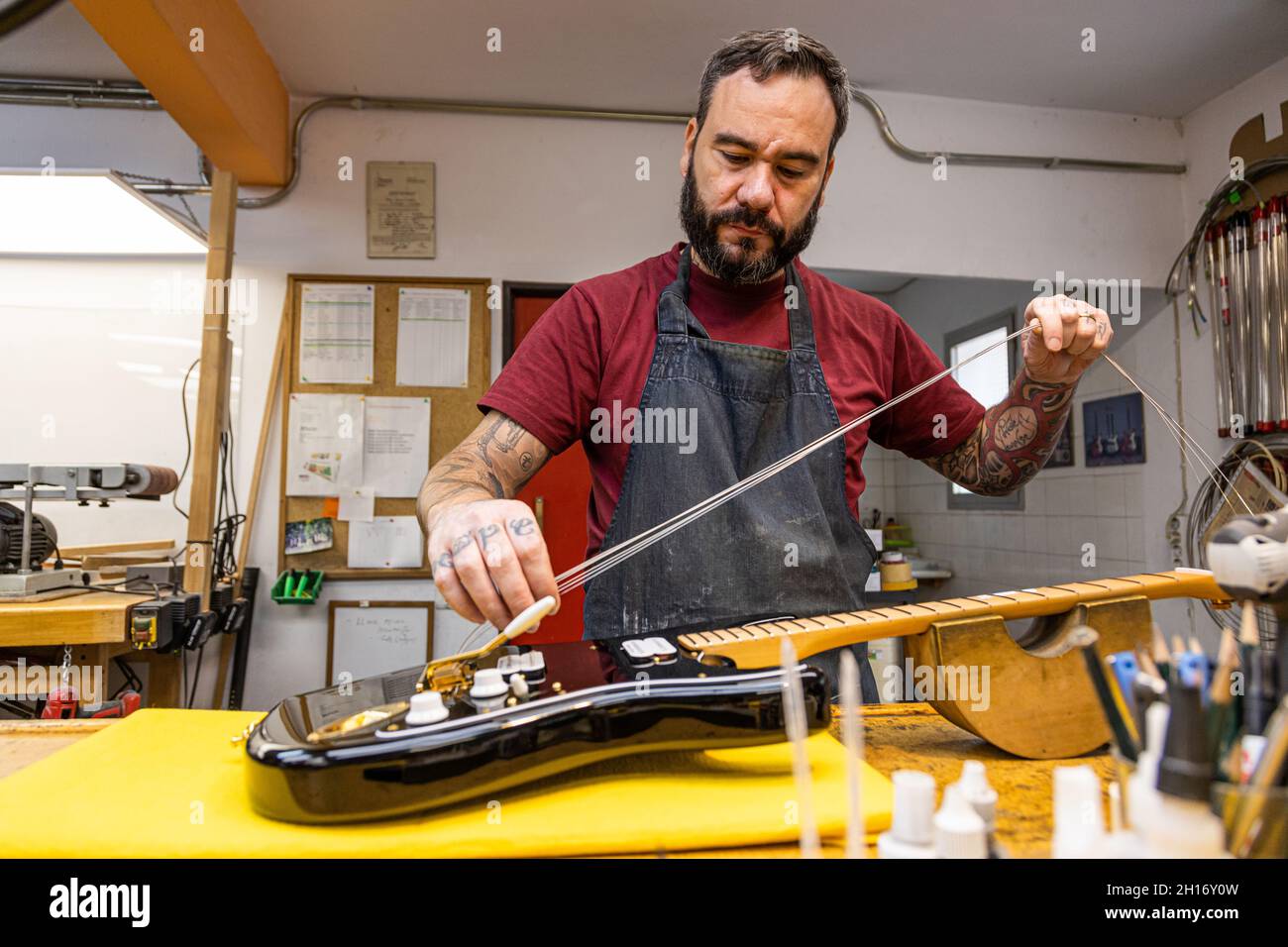 Artisan qualifié en tablier debout et changeant de cordes sur guitare électrique dans un atelier professionnel Banque D'Images