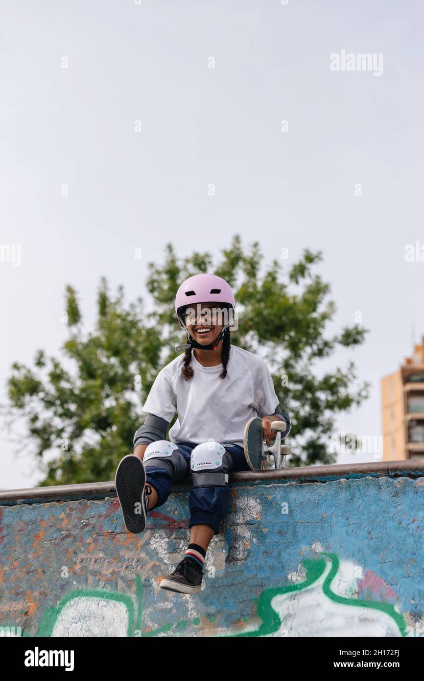 Corps complet de jeune afro-américaine gaie portant un t-shirt blanc avec casque et des coussins de sécurité avec planche à roulettes assis sur la rampe de skate dans le parc Banque D'Images