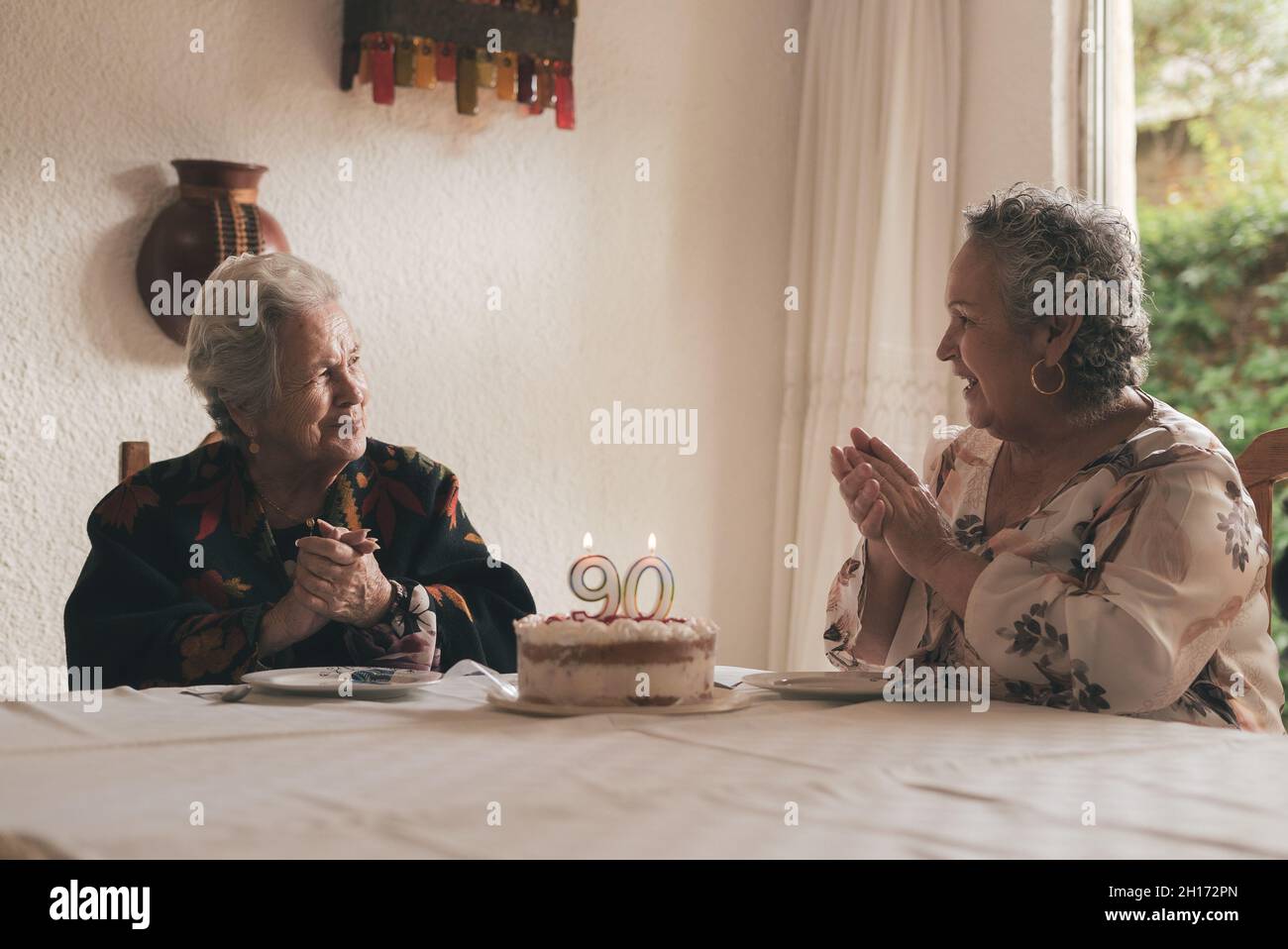 Femme âgée aux cheveux gris et femme sénior assis à la table à manger et célébrant l'anniversaire de 90th avec un délicieux gâteau aux bougies Banque D'Images