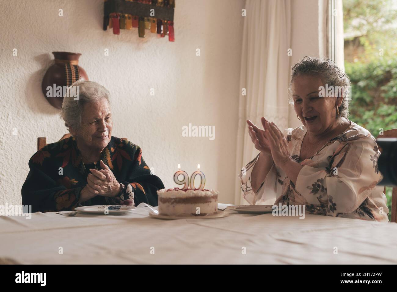 Femme âgée aux cheveux gris et femme sénior assis à la table à manger et célébrant l'anniversaire de 90th avec un délicieux gâteau aux bougies Banque D'Images