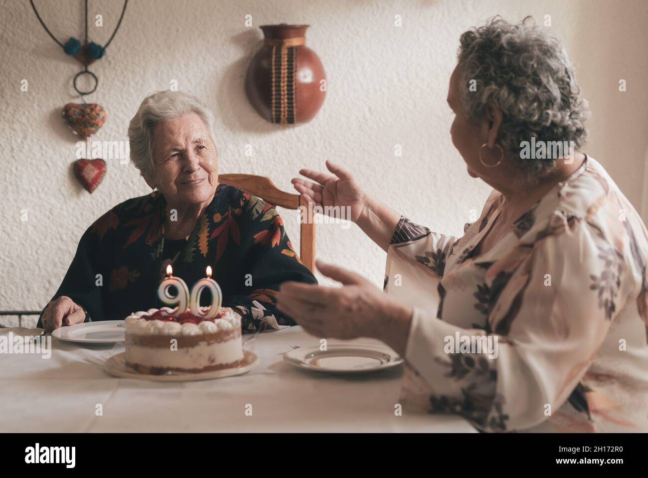 Femme âgée aux cheveux gris et femme sénior assis à la table à manger et célébrant l'anniversaire de 90th avec un délicieux gâteau aux bougies Banque D'Images