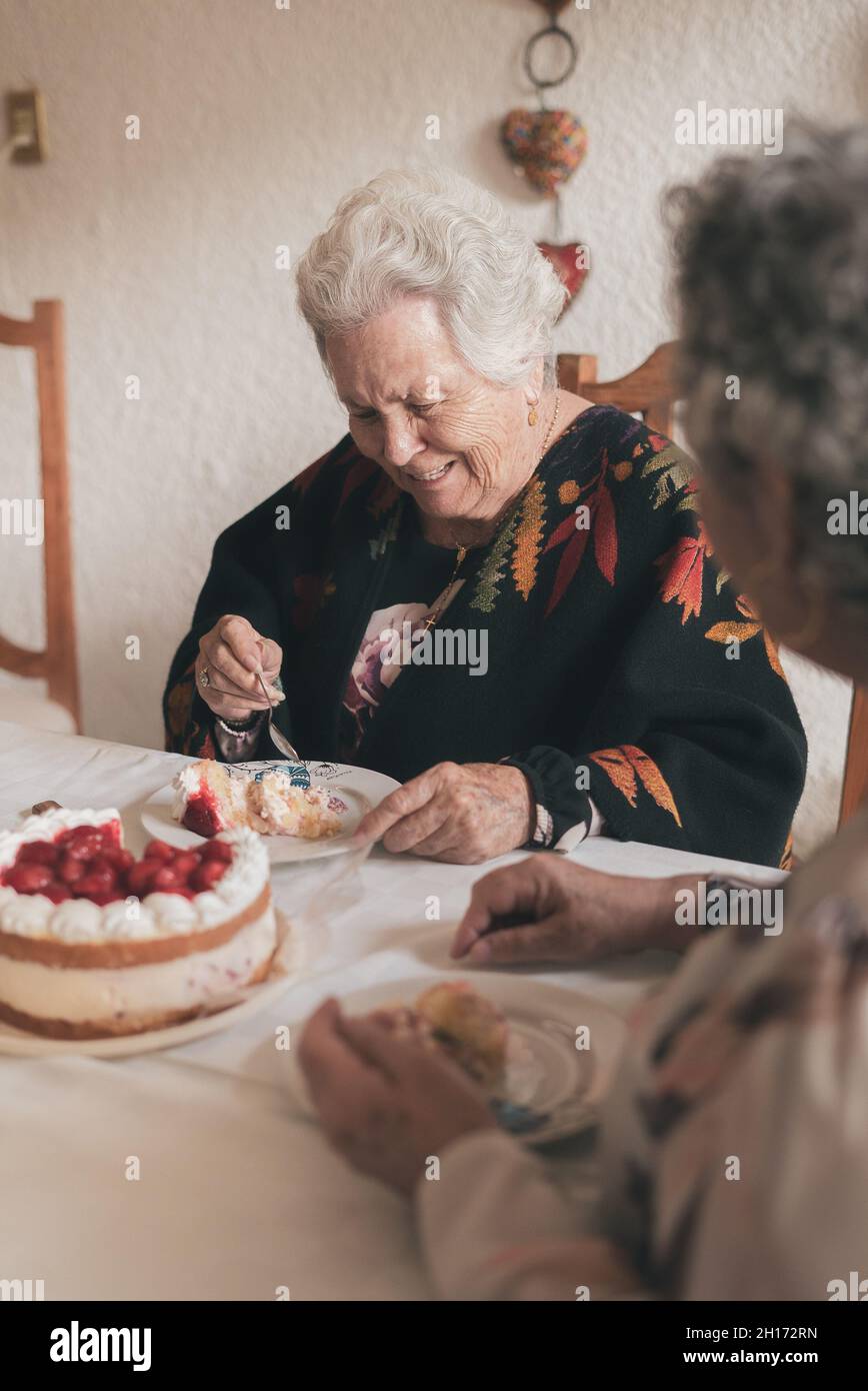 Femme âgée aux cheveux gris et femme sénior assis à la table à manger et célébrant l'anniversaire de 90th avec un délicieux gâteau aux bougies Banque D'Images