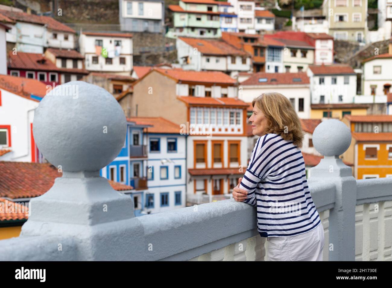 Vue latérale d'une femme de voyage mûre penchée sur la clôture tout en admirant la vue sur les maisons résidentielles colorées typiques de la ville des Asturies Banque D'Images