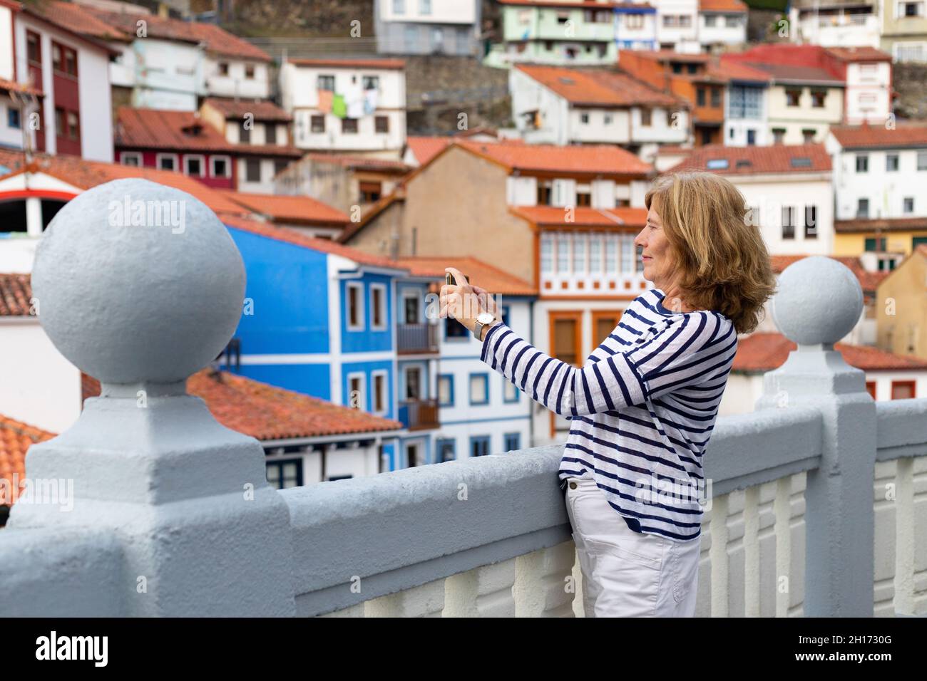 Vue latérale d'une femme voyageur âgée prenant des photos de maisons résidentielles miteuses sur un smartphone près de la clôture pendant le voyage dans les Asturies Banque D'Images