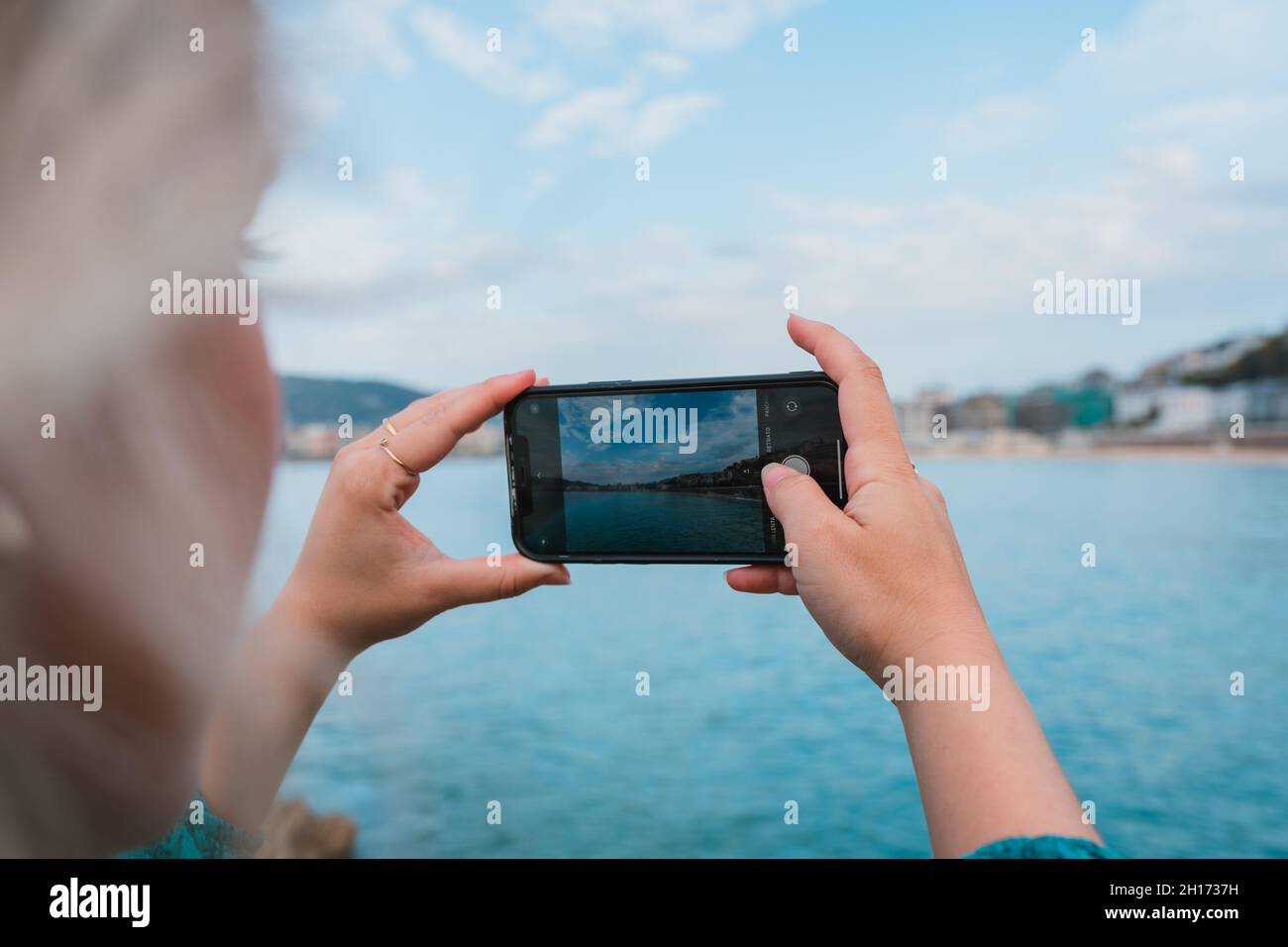 Vue latérale d'une femme élégante et méconnaissable aux cheveux blonds prenant en photo l'océan bleu contre le paysage urbain de Donostia sur un smartphone en journée Banque D'Images