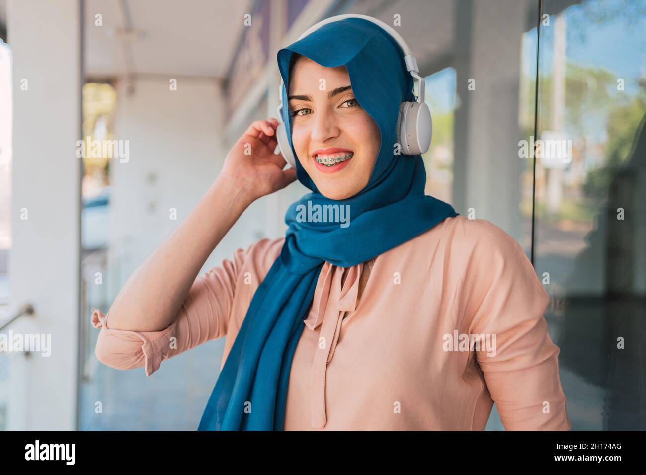 Femme arabe positive en foulard et casque à l'écoute de musique regardant  l'appareil photo et appréciant des chansons tout en se tenant dans la rue  de la ville Photo Stock - Alamy