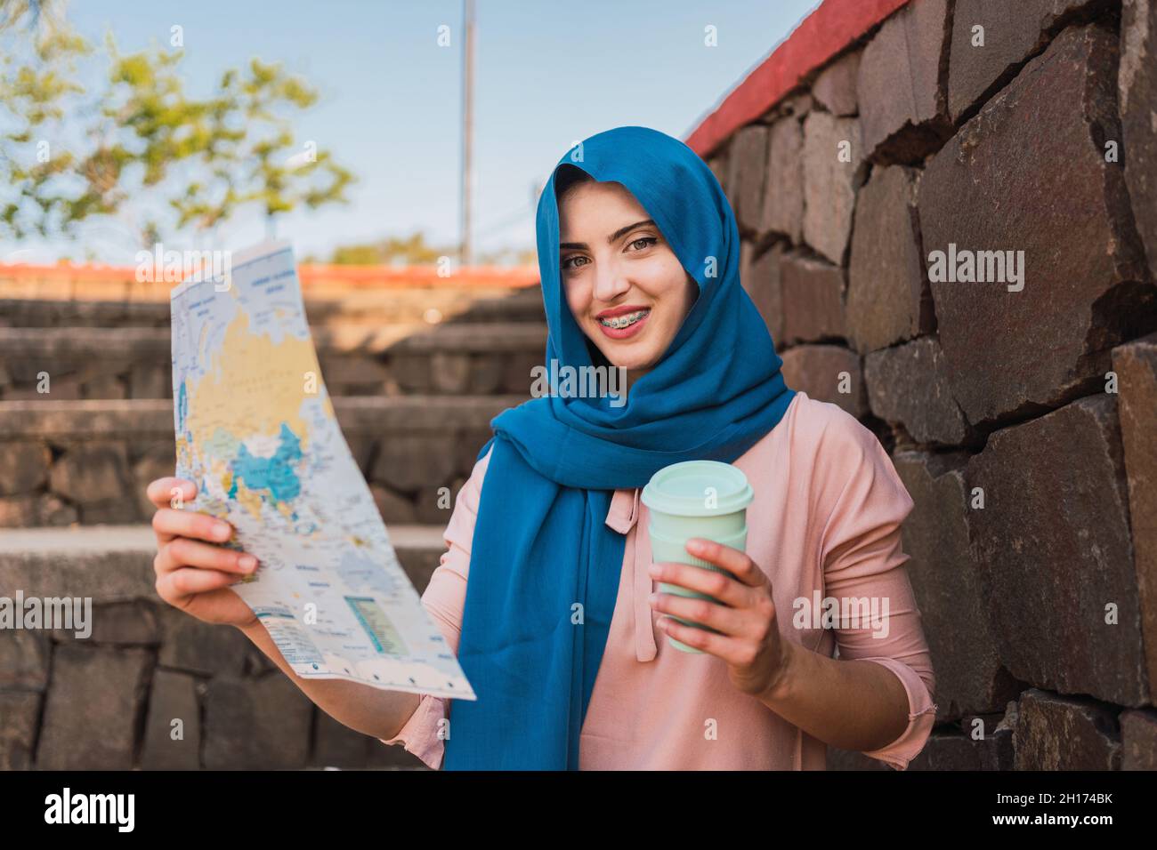 Femme musulmane concentrée dans un foulard et avec un café à emporter  lisant la carte sur papier et recherchant la direction tout en se tenant  près du mur de pierre Photo Stock -