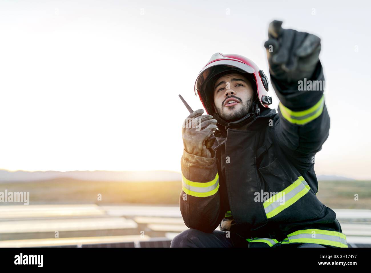 Pompier positif, jeune homme barbu, portant un casque et des gants et des vêtements de protection tout en tenant le talkie-walkie dans la main levée et paisible Banque D'Images