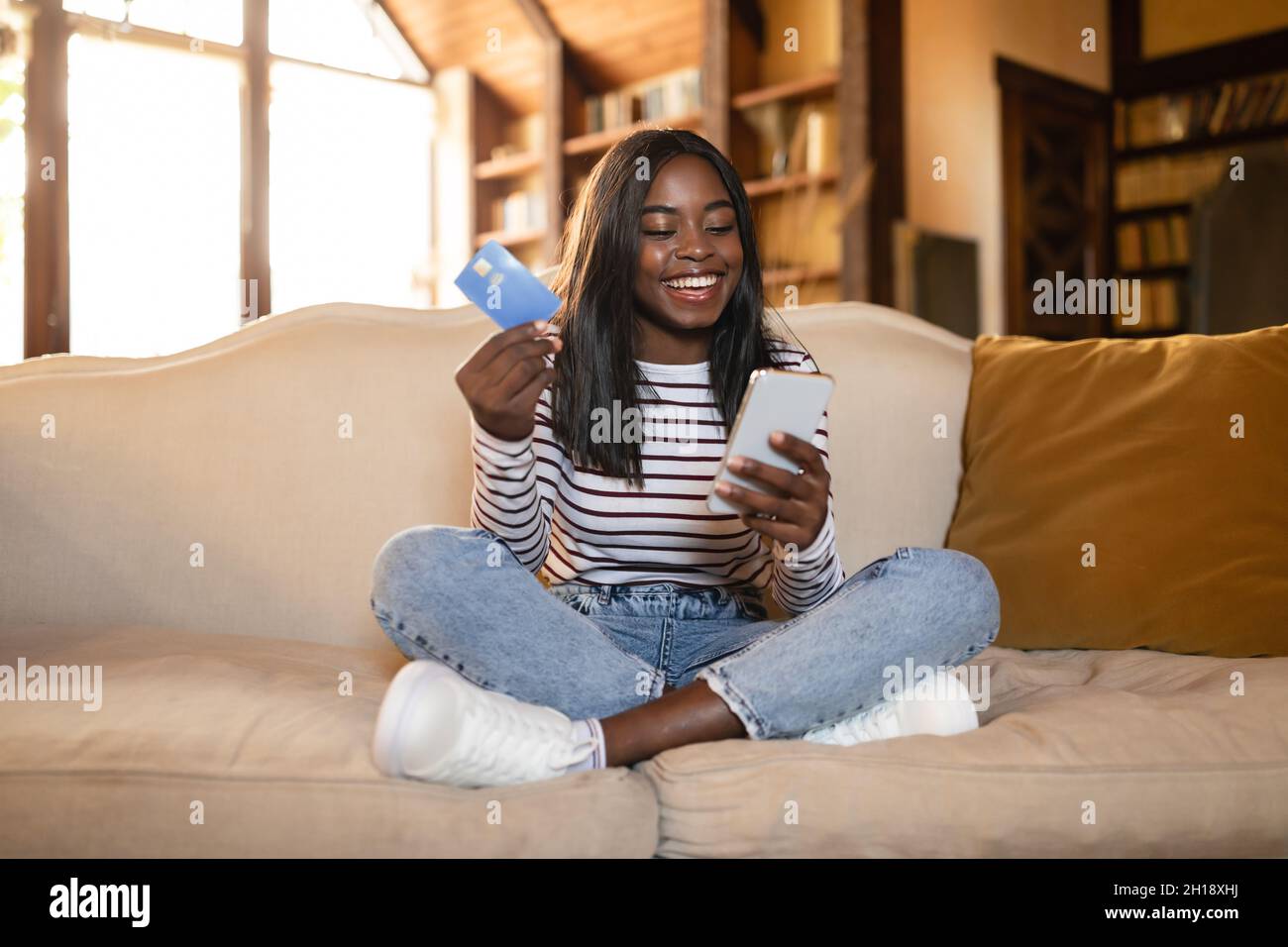 Pleine longueur de jeune femme noire avec carte de crédit et smartphone shopping dans la boutique en ligne, assis sur un canapé à la maison Banque D'Images
