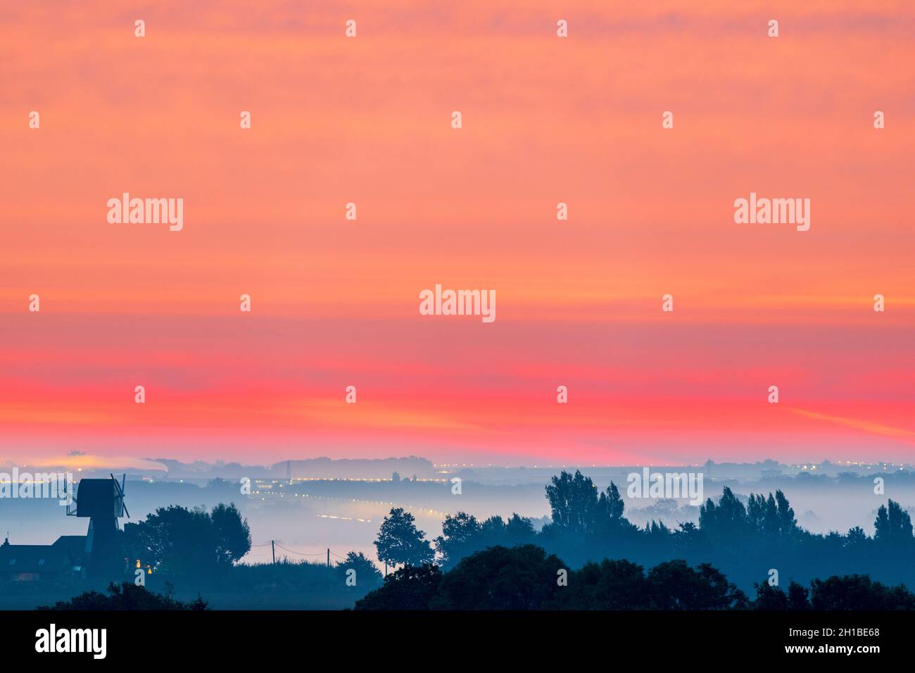 Le ciel d'aube rouge au-dessus d'une brume couvrait le Kent Paysage avec quelques arbres et un ancien moulin à vent silhoueté contre la brume de mer.Le ciel est rouge et des bandes orange sont horizontales.Horizon bas dans le cadre. Banque D'Images