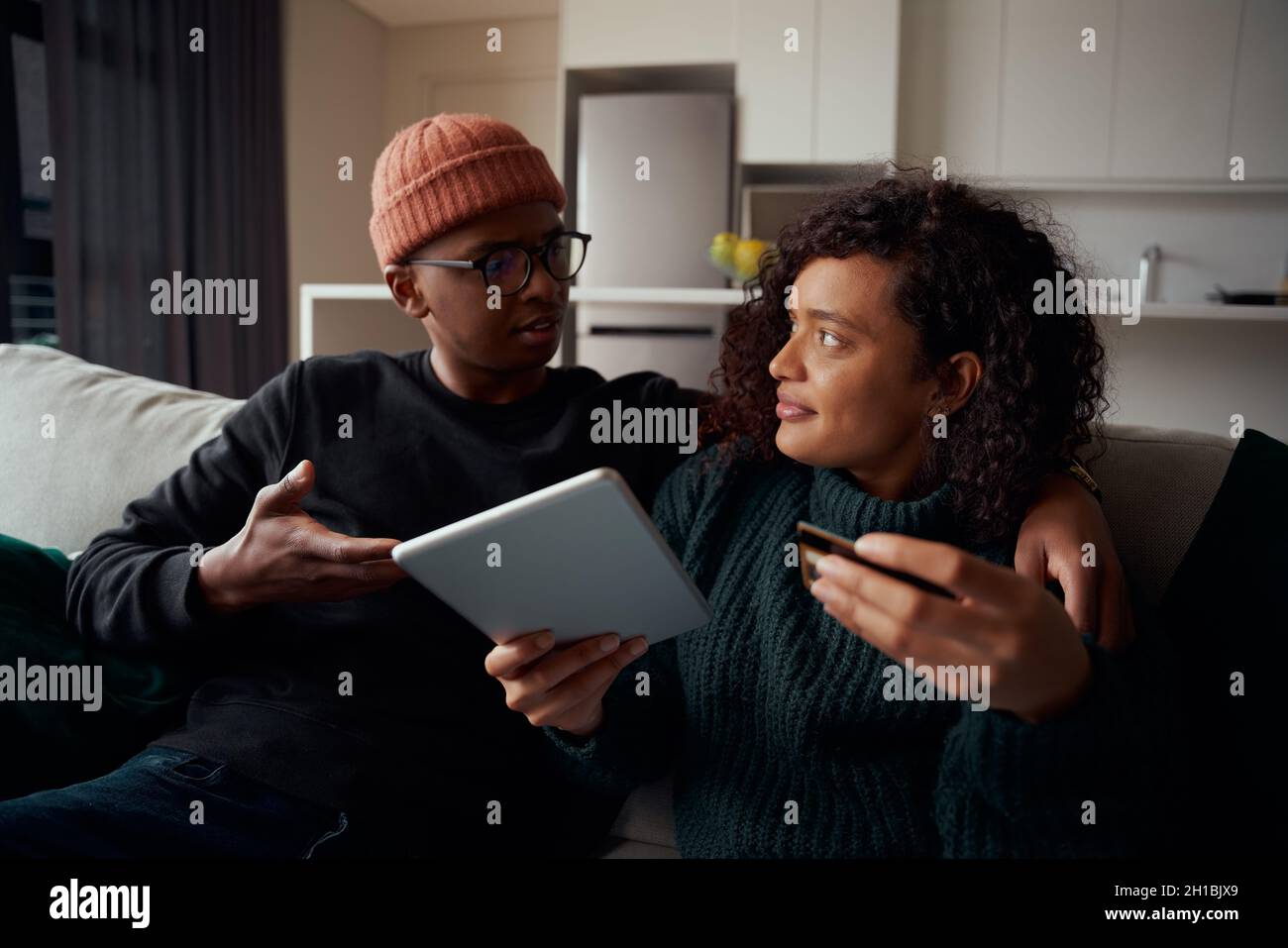 Un couple multiethnique se bat pour acheter des produits en ligne sur une tablette avec une carte de crédit, assis sur un canapé dans le salon Banque D'Images