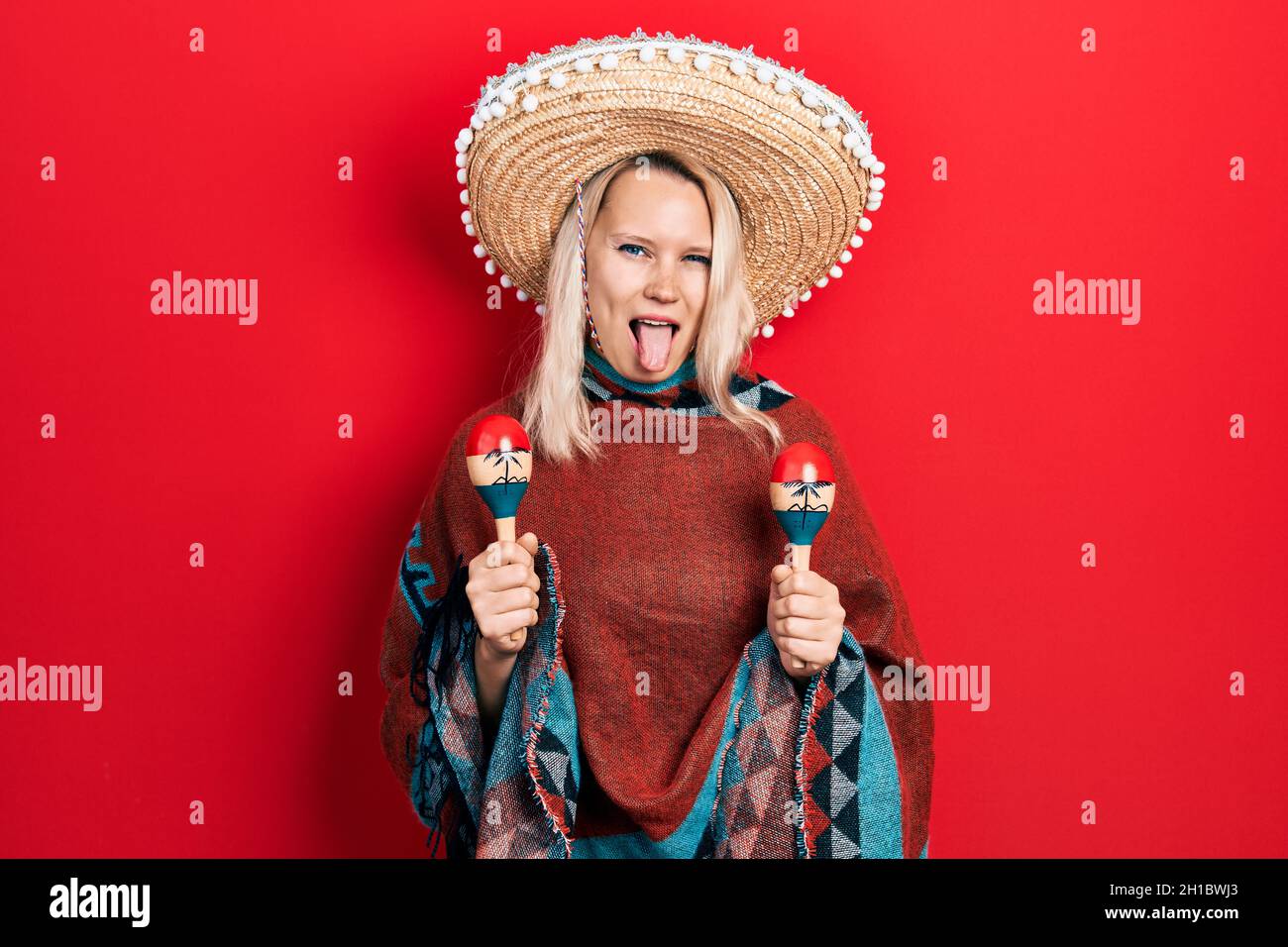 Belle femme blonde caucasienne portant un poncho mexicain festif et des maracas qui collent la langue heureux avec l'expression drôle. Banque D'Images