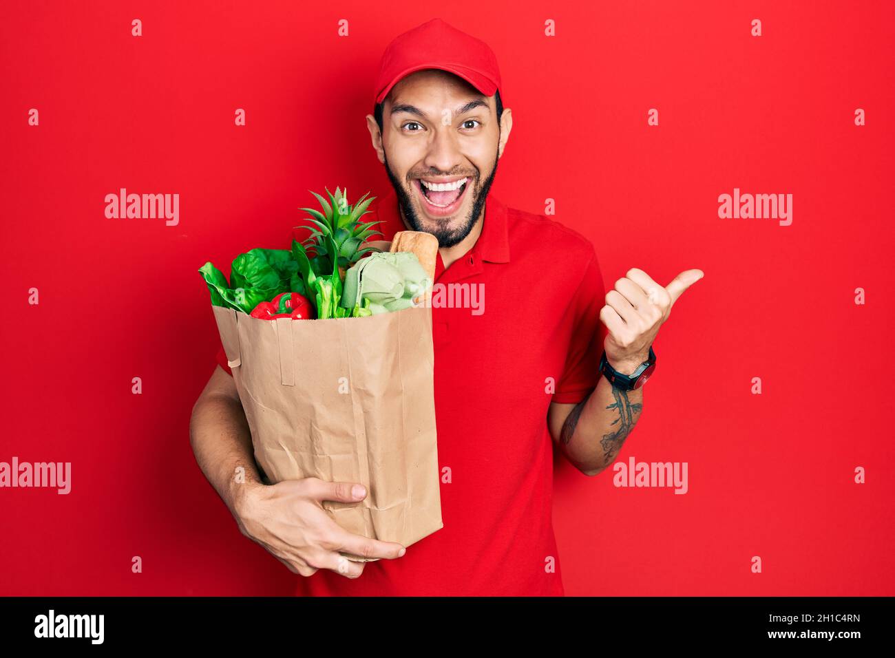 Homme hispanique avec barbe portant l'uniforme de messagerie avec des provisions du supermarché pointant le pouce vers le côté souriant heureux avec la bouche ouverte Banque D'Images