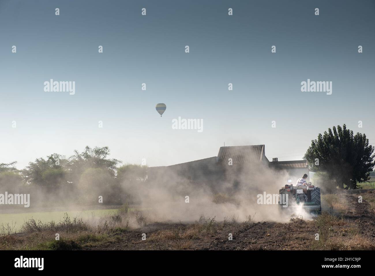 un jeune agriculteur travaillant dans le champ avec un tracteur équipé d'un ballon d'air chaud qui traverse le ciel Banque D'Images