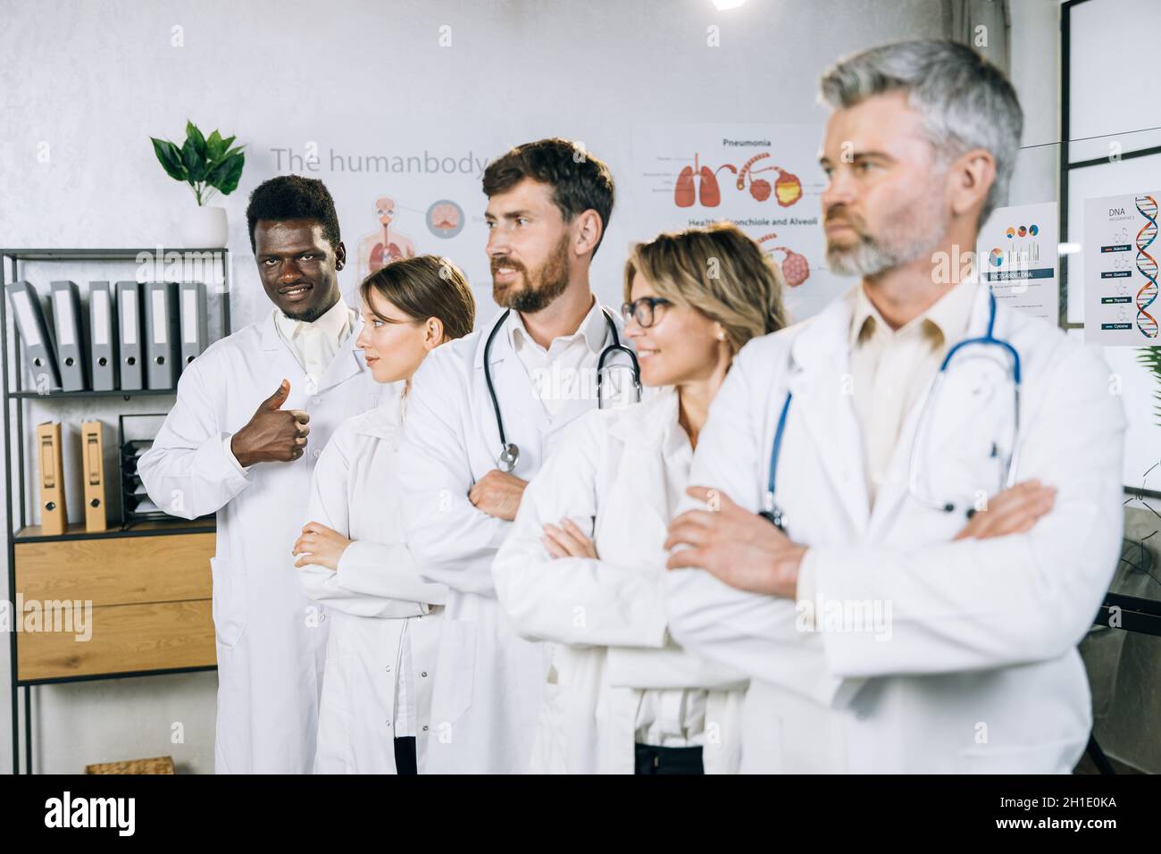 Un homme afro-américain se tenant en ligne avec des collègues multiraciaux, souriant et montrant le pouce sur la caméra.Équipe de spécialistes médicaux se posant dans un cabinet moderne. Banque D'Images