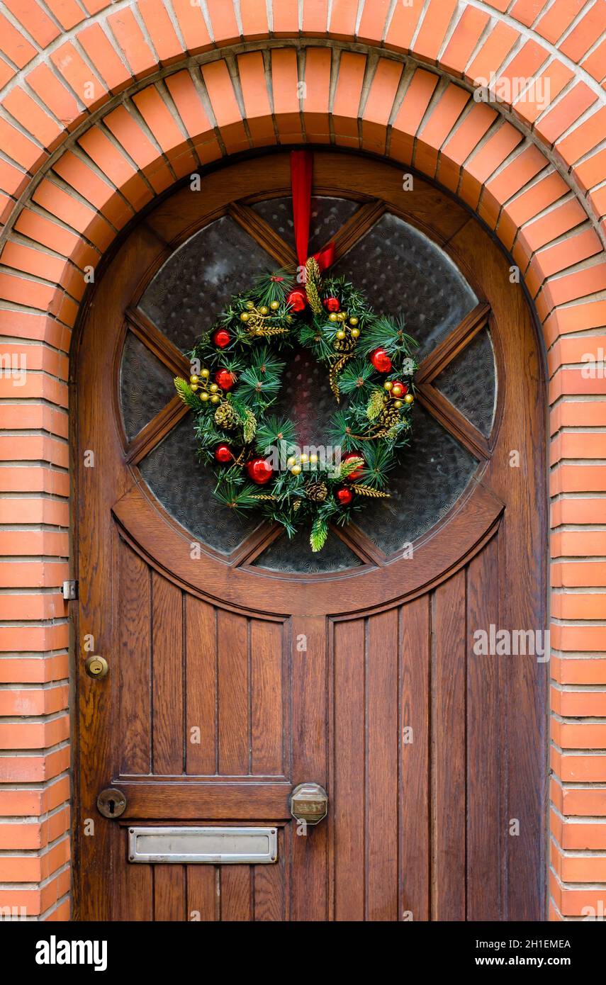 Vue avant d'une couronne de Noël en branches de sapin en plastique avec boules et cônes de pin, accrochée sur une porte en bois à moitié vitrée avec une fram en brique Banque D'Images