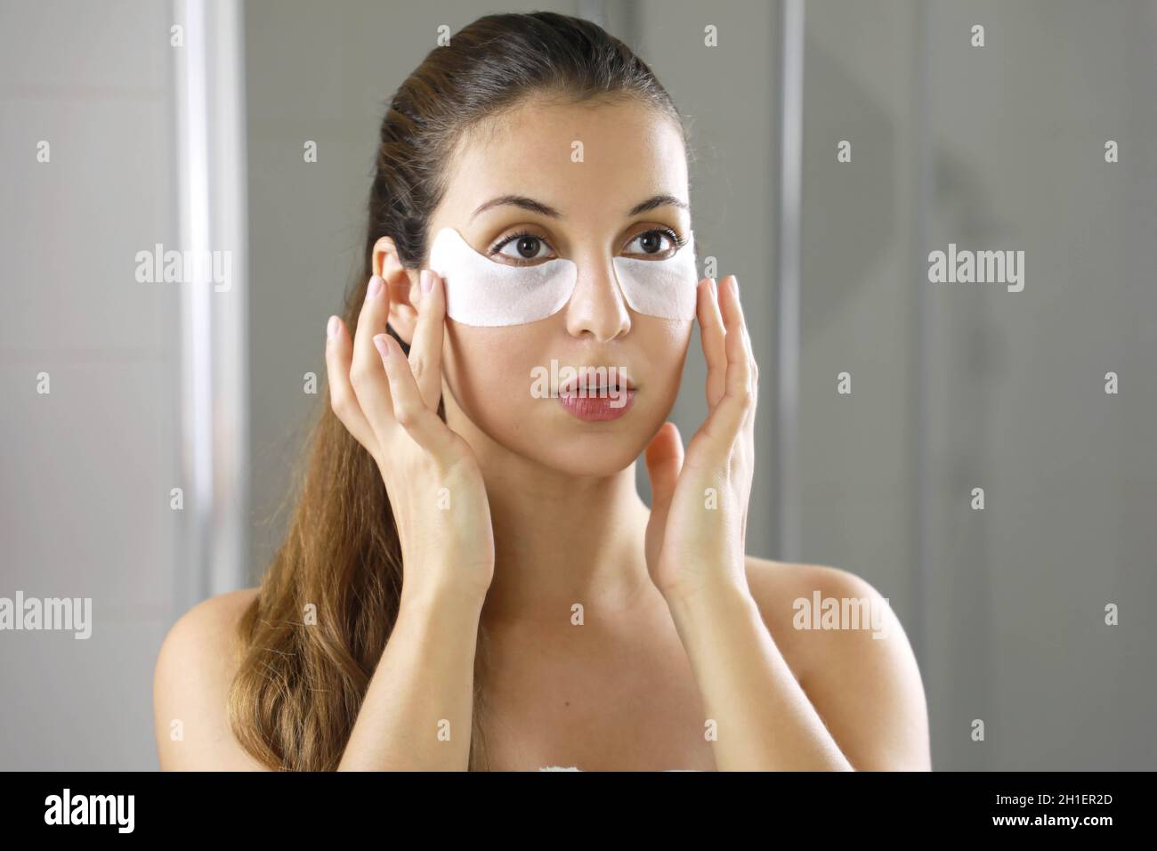 Femme de beauté appliquant le masque sous-oculaire regardant elle-même dans le miroir dans la salle de bains. Soins de la peau fille toucher des patches de tissu masque sous les yeux pour réduire e Banque D'Images