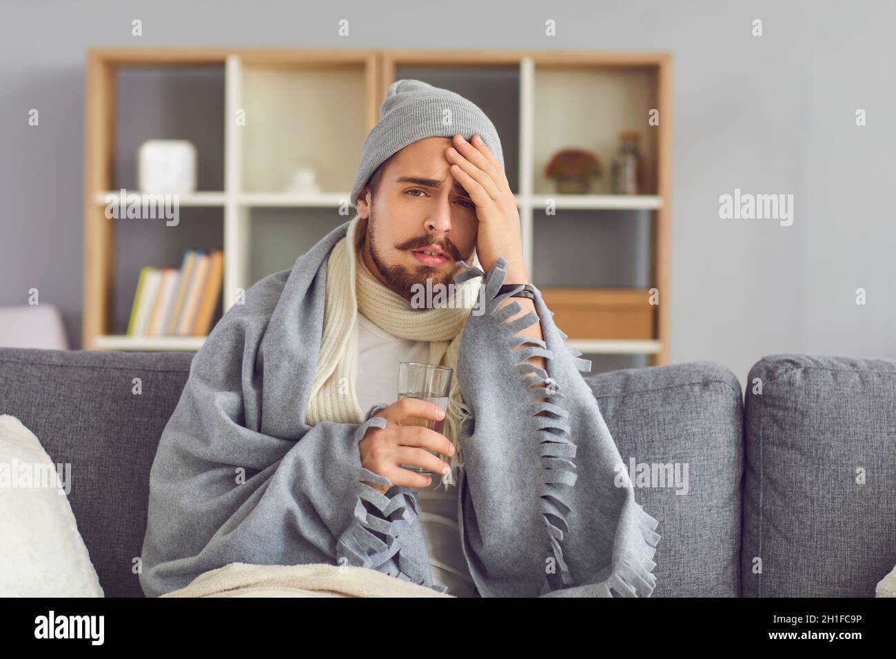 Un homme malade avec un verre d'eau dans sa main souffre d'un grand mal de tête assis sur le canapé. Banque D'Images