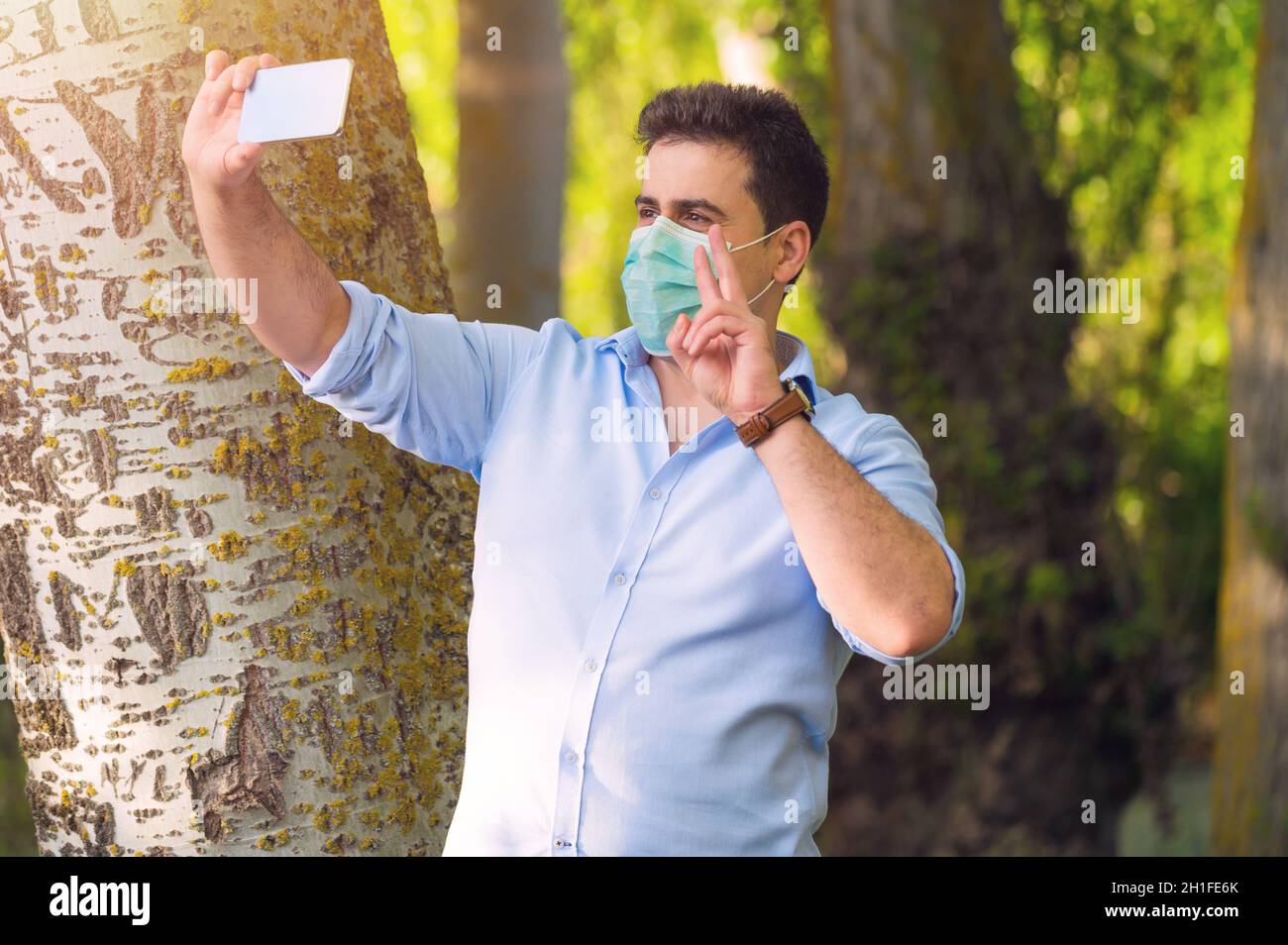 COVID-19 beau homme optimiste portant un masque chirurgical sur le visage pendant la maladie pandémique du coronavirus montrant un signe de victoire dans le parc de la ville tout en faisant un vid Banque D'Images