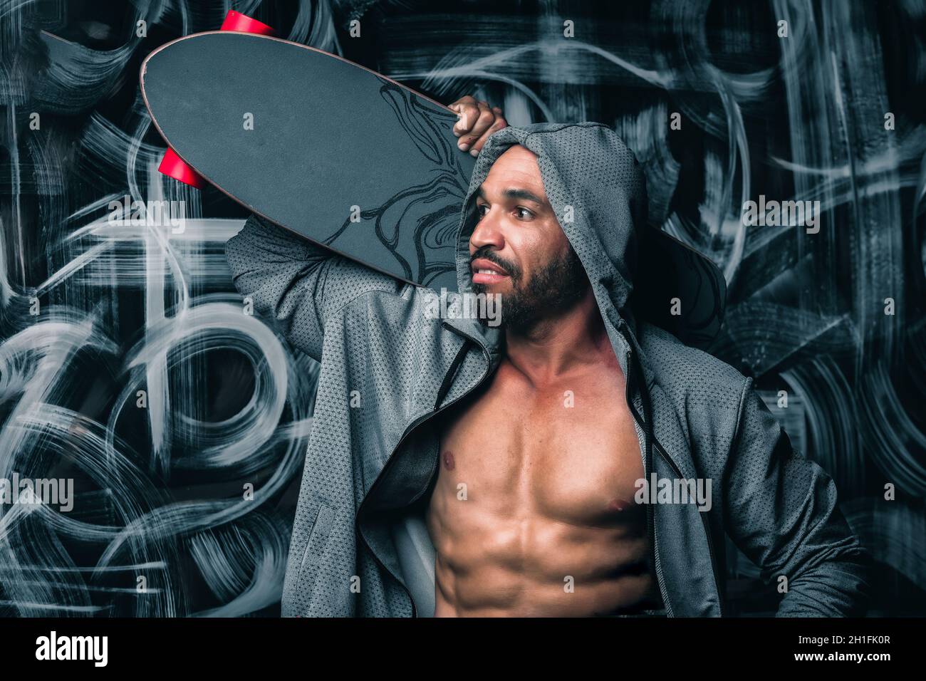Un Latino hivers dans une veste de sport se tient contre le mur et tient un skateboard sur son épaule.Portrait d'un homme avec une longue planche.Style urbain. Banque D'Images