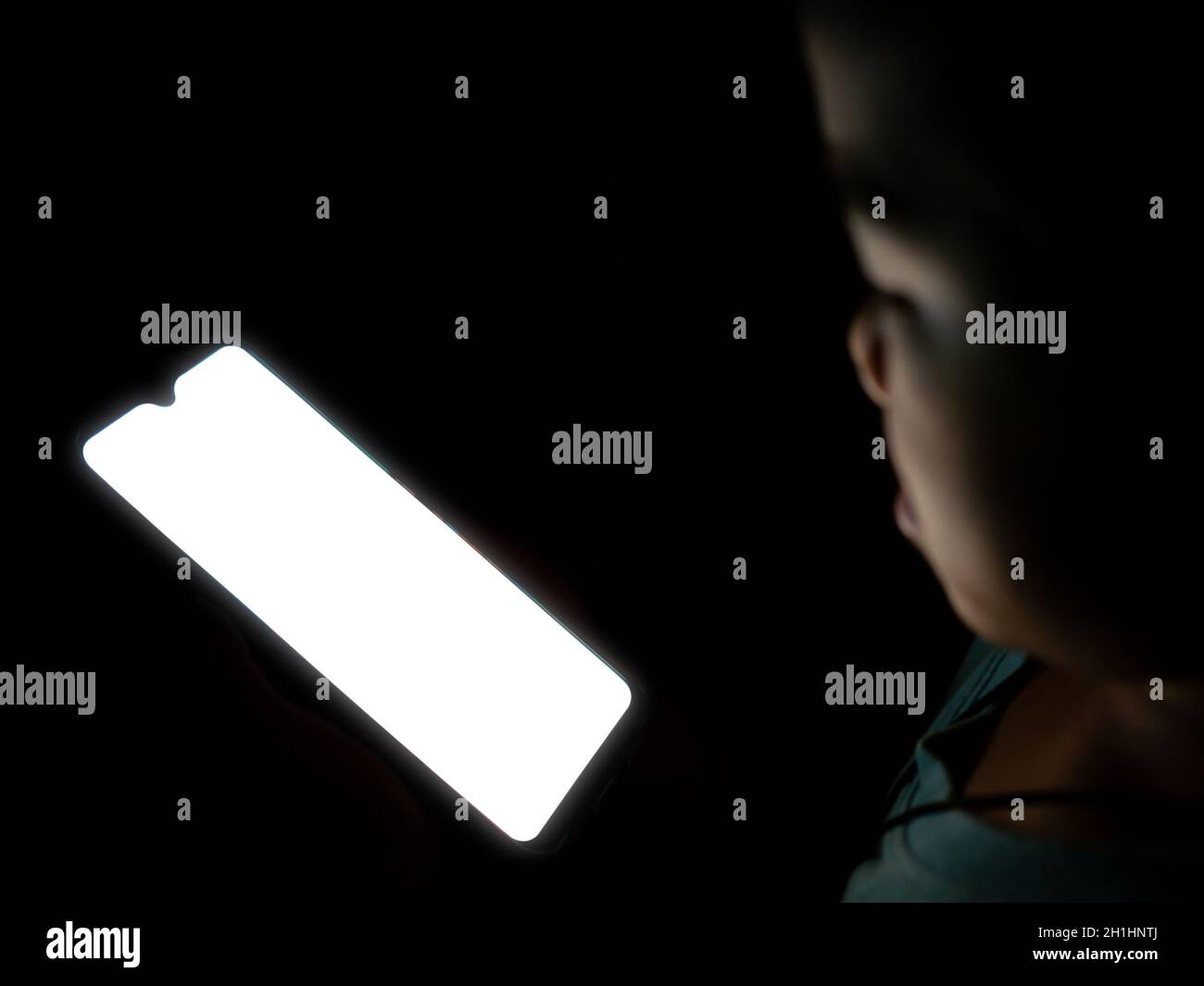 Un garçon regardant un écran blanc de téléphone blanc sur un fond noir foncé. Banque D'Images