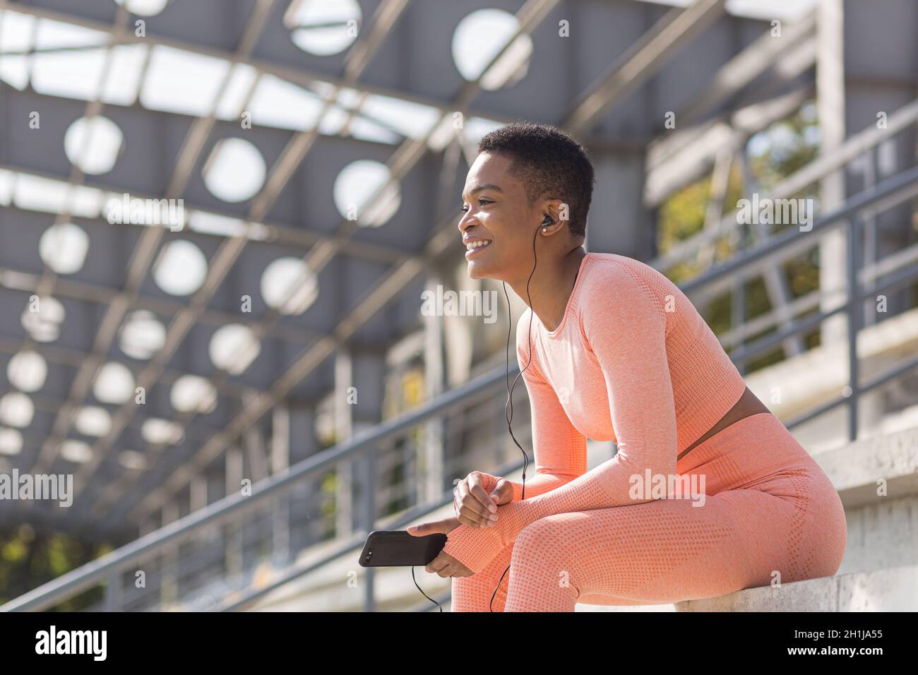 Belle et réussie athlète féminine souriante écoute le livre audio de téléphone mobile pendant la forme physique et le jogging du matin près du stade, african ameri Banque D'Images