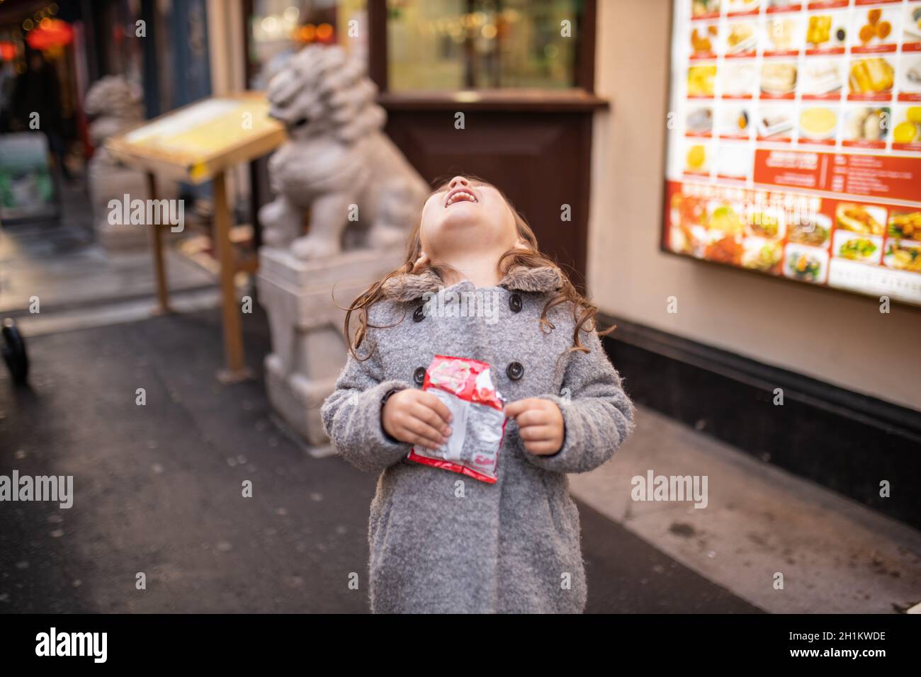 La photo d'une petite fille brunette en manteau gris regarde comme elle rit hystériquement, et avec un restaurant chinois flou et la statue d'un traditi Banque D'Images