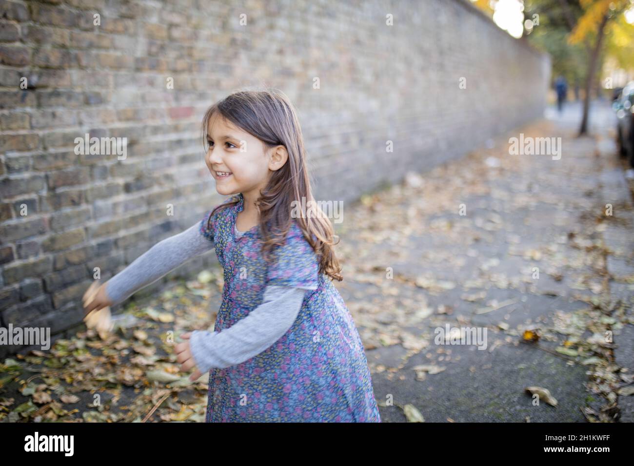 Photo d'une petite fille brune heureuse et vibrante portant une robe fleurie sur le trottoir recouvert de feuilles d'automne et à côté d'un mur de briques Banque D'Images