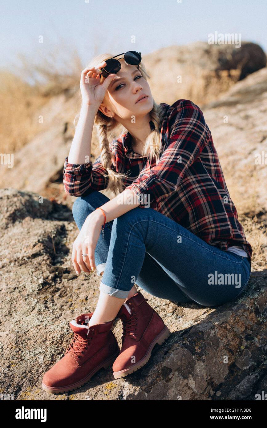 fille blonde avec des queues de porc dans une chemise, un jean, des chaussures rouges et des verres noirs sur des rochers de granit Banque D'Images