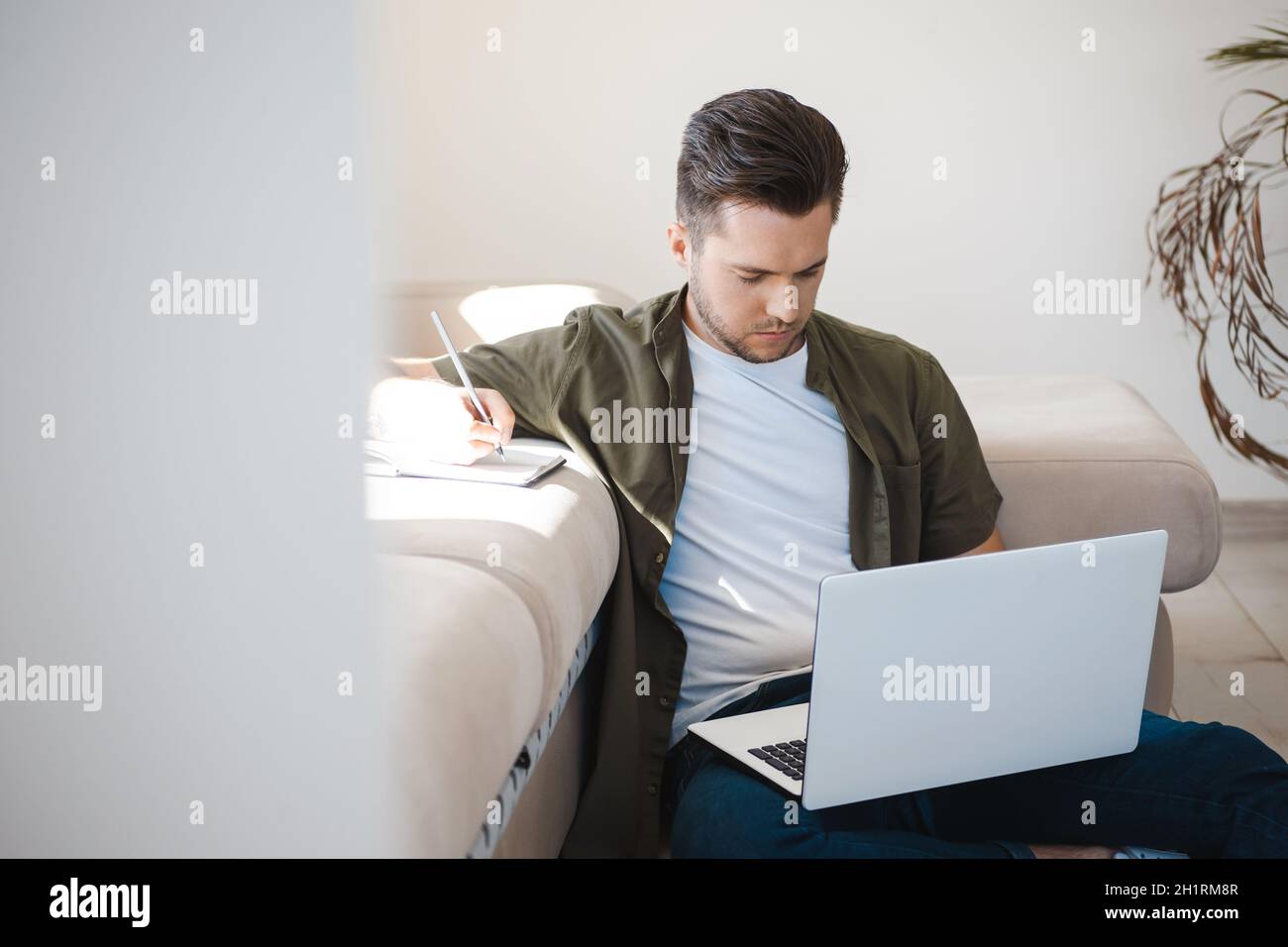 Homme participant à une conférence en ligne assis sur le sol et prenant des notes dans son carnet.Regarder vers le bas Banque D'Images