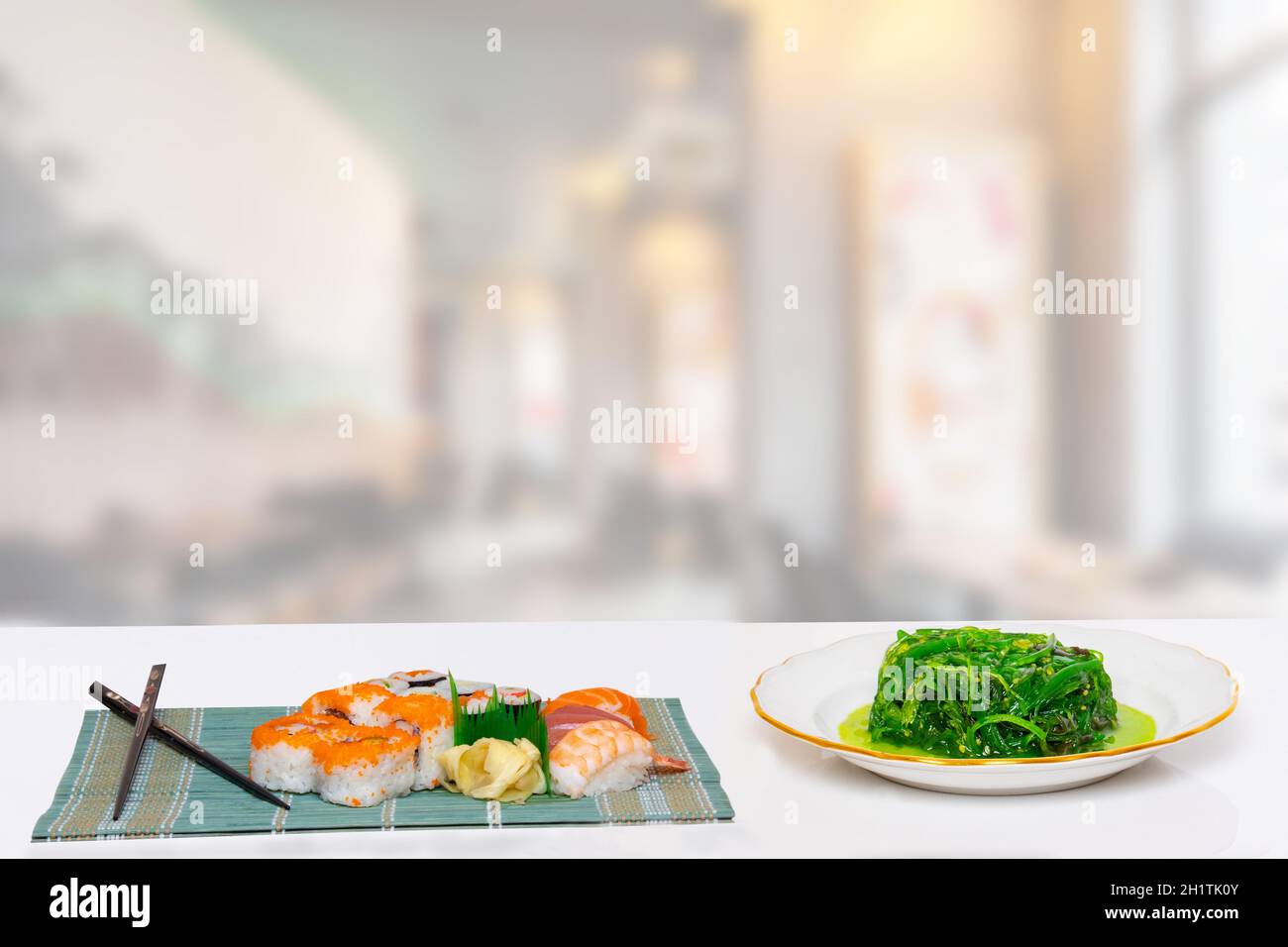 Gros plan de divers types de sushis et de baguettes servis tapis de bambou vert et salade de chuka fraîche avec sesam plaque sur une table blanche sur une ja floue Banque D'Images