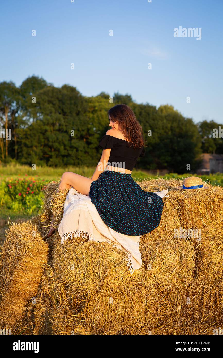 Jeune belle femme dans un chapeau de paille regarde le soleil couchant, la fille est assise sur une grande pile de balles de paille. Chaude soirée d'été Banque D'Images