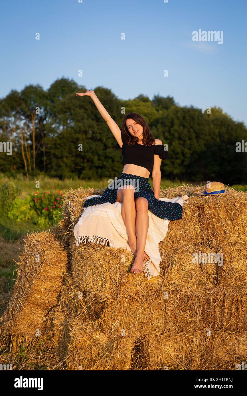 Jeune belle femme regarde le soleil couchant et fait des vagues sa main, s'assoit sur une grosse pile de balles de paille Banque D'Images