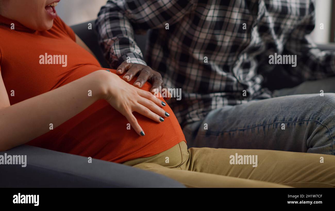 Gros plan des mains interraciales du jeune couple sur le ventre en attendant l'enfant tout en étant assis sur le canapé.Les personnes multiethniques touchant la bosse de bébé se sentent gaies et parlent de la parentalité Banque D'Images