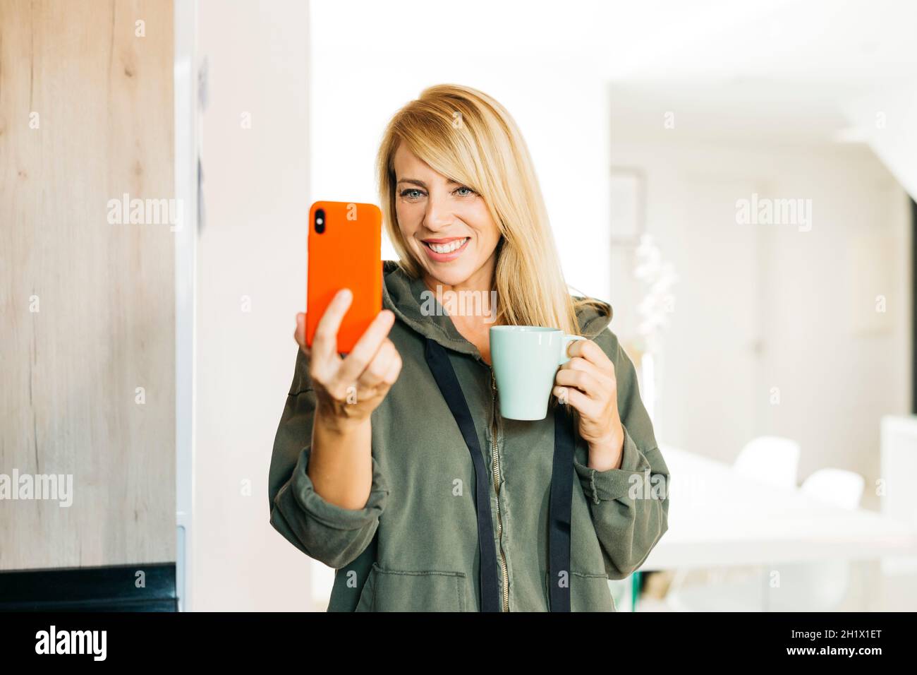 Femme blonde d'âge moyen buvant du café et ayant un appel vidéo dans une cuisine Banque D'Images