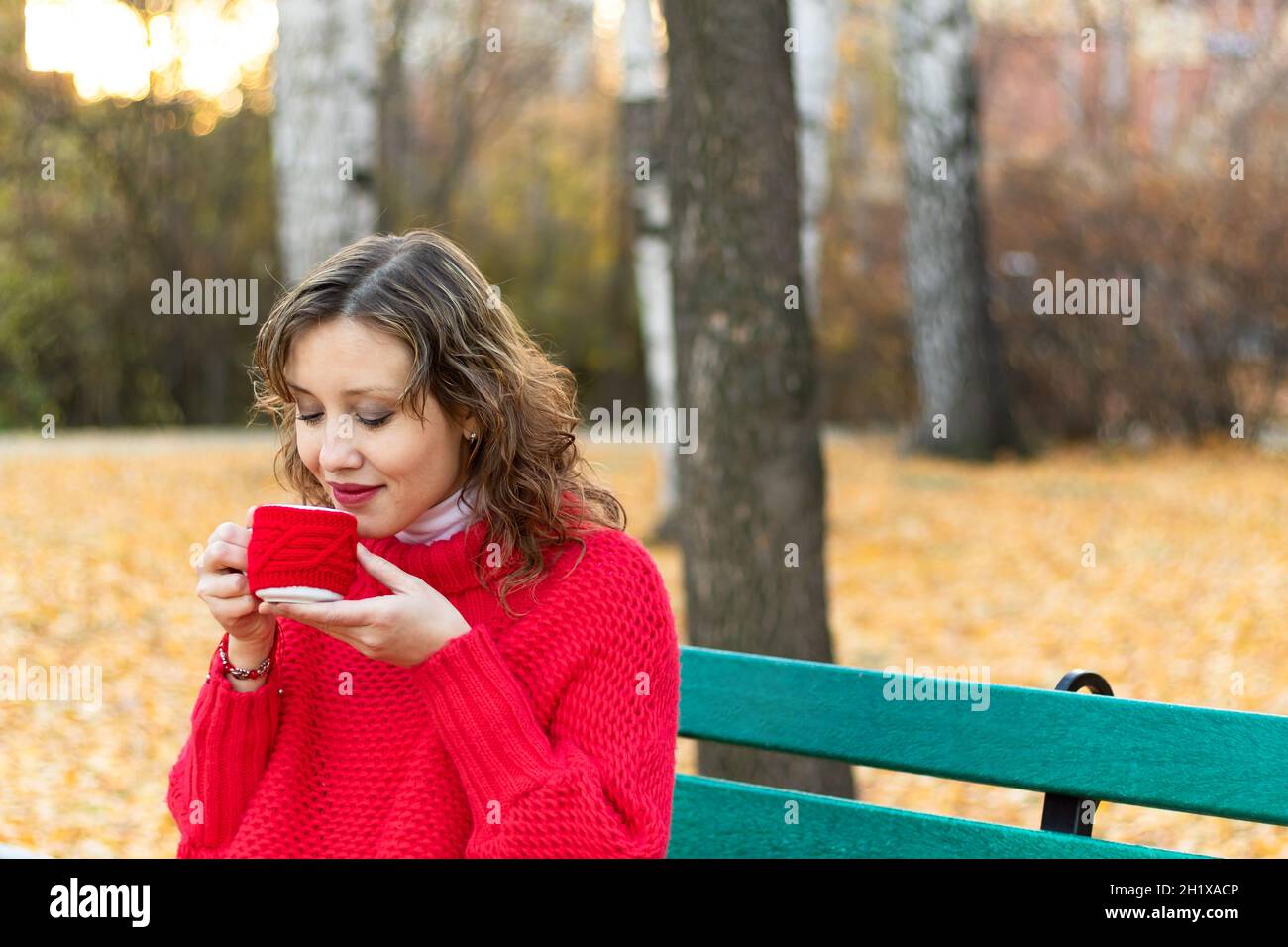 Une belle jeune femme dans un chandail et un bracelet rouge vif est assise sur un banc de parc et tient une tasse rouge avec une boisson chaude dans ses mains contre le dos Banque D'Images