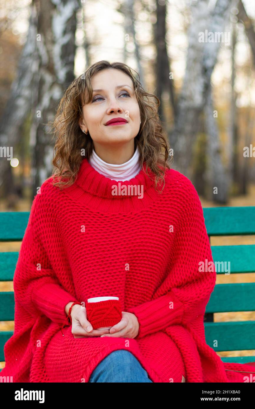 Une belle jeune femme dans un chandail et un bracelet rouge vif est assise sur un banc de parc et tient une tasse rouge avec une boisson chaude dans ses mains contre le dos Banque D'Images