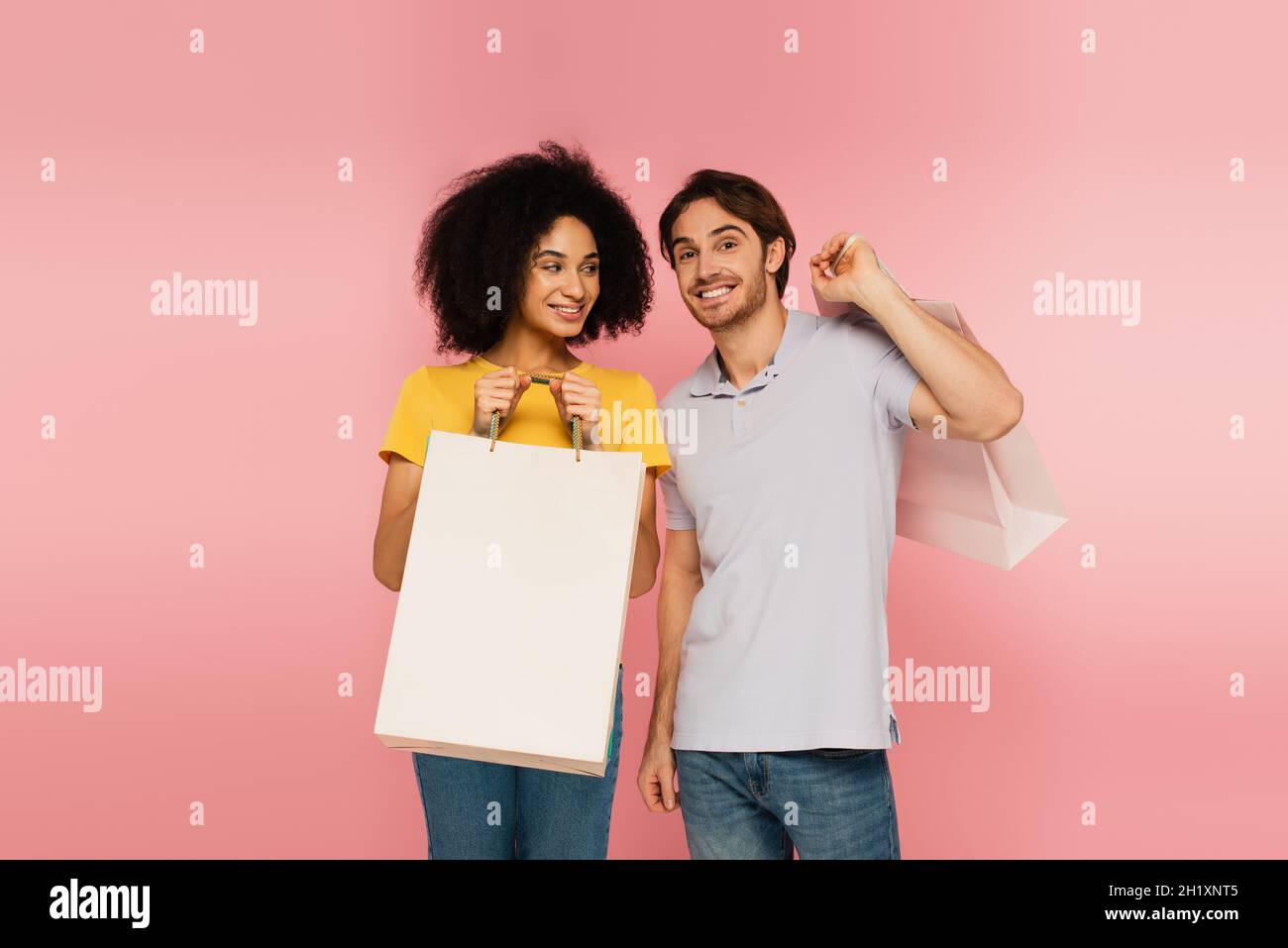 homme joyeux souriant à l'appareil photo près d'une femme hispanique avec sac de shopping isolé sur rose Banque D'Images