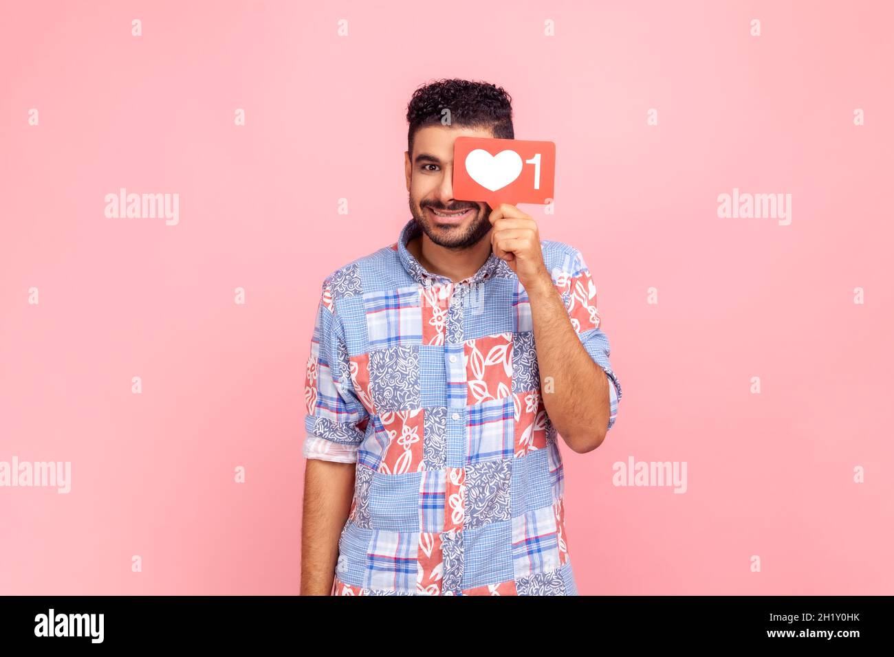 Homme souriant en bleu décontracté chemise cache le visage derrière le comptoir rouge aime, regarde la caméra avec un sourire crasseux, la gestion smm dans les réseaux sociaux.Studio d'intérieur isolé sur fond rose. Banque D'Images