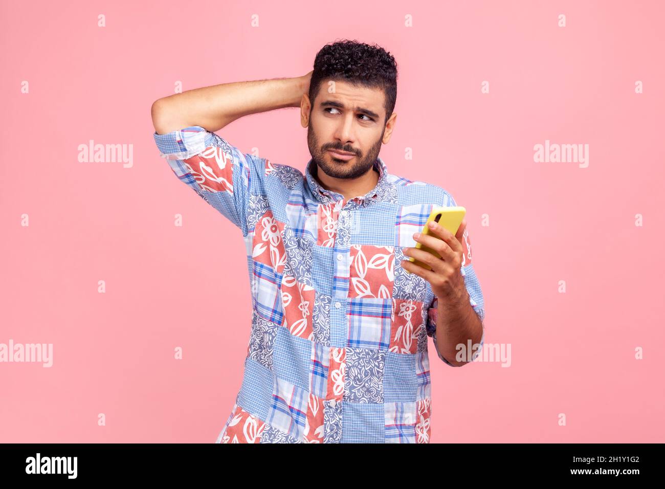 Homme barbu réfléchi en chemise bleue à la tête égratigante tenant le smartphone à la main, en envisageant la mise à jour de logiciels, en choisissant des tarifs appropriés.Studio d'intérieur isolé sur fond rose. Banque D'Images