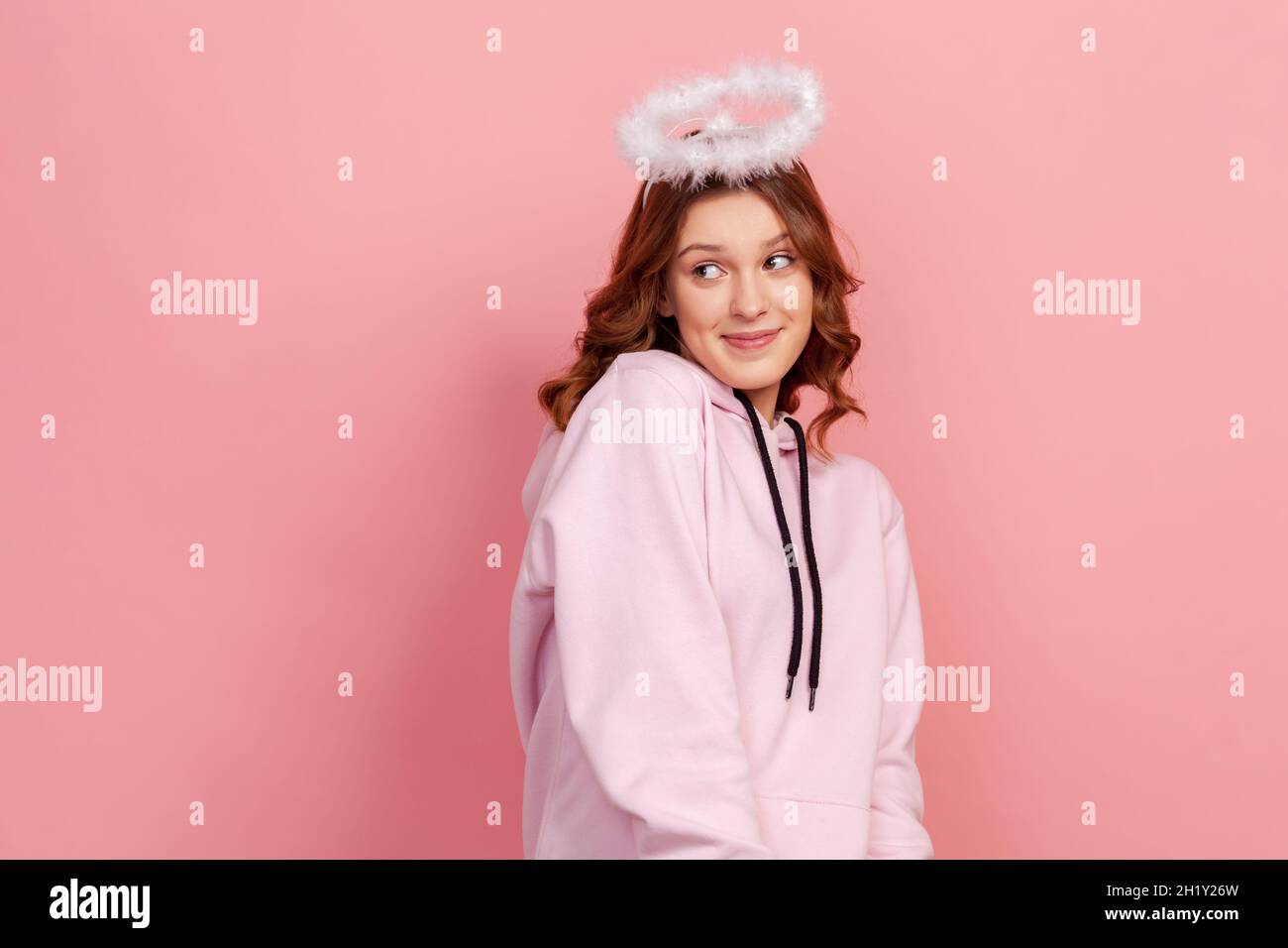 Portrait d'une jeune fille à poil dur timide debout avec un halo sur sa tête et regardant loin avec le sourire et le visage rêvant, ange.Studio d'intérieur isolé sur fond rose Banque D'Images